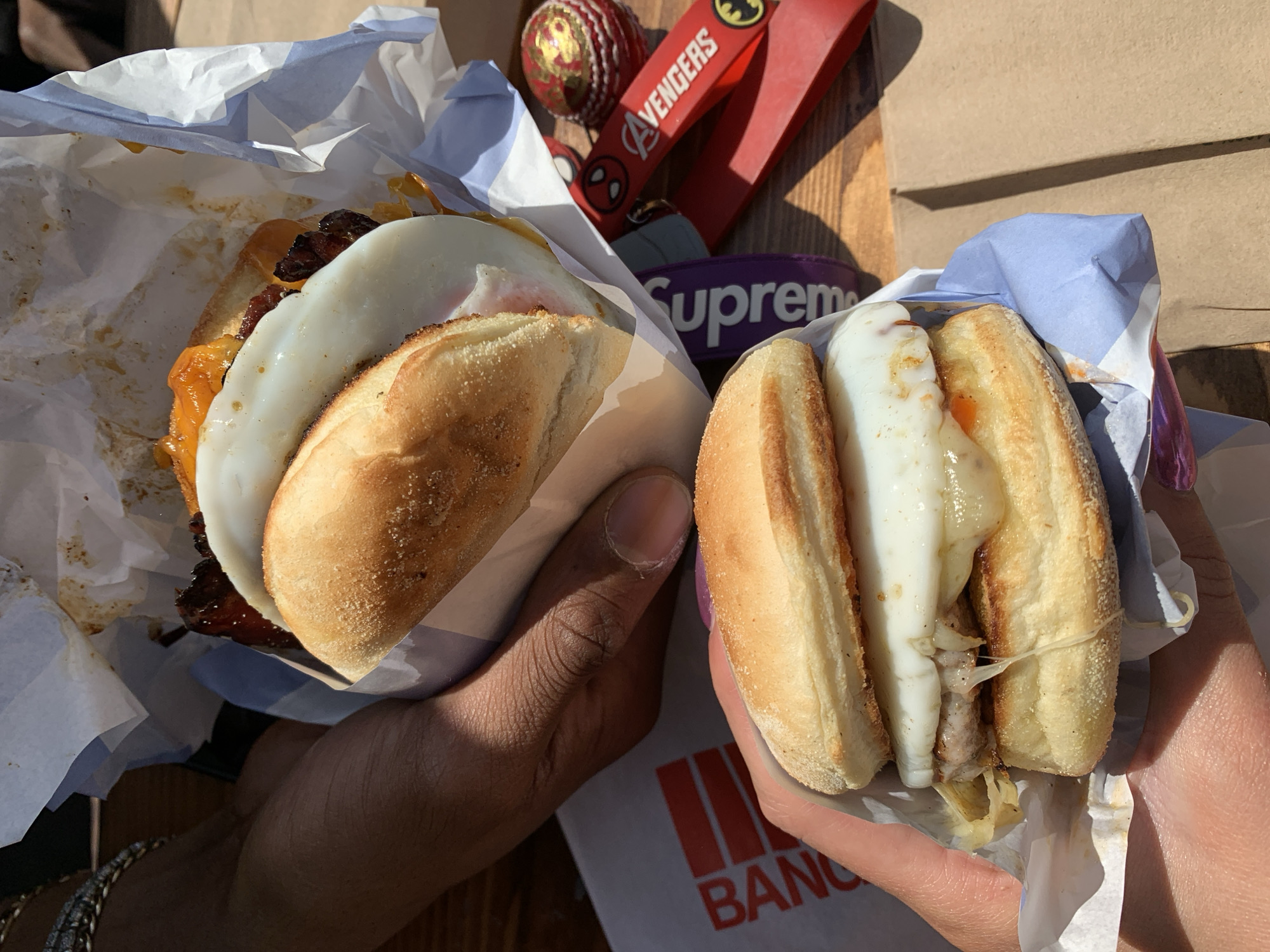 This bright, high-angle image captures two hands holding breakfast sandwiches outdoors, against the backdrop of what is identified as London, United Kingdom. The clear, sunny weather is evident from the strong sunlight casting distinct shadows.
On the left, a hand with darker skin holds a sandwich made with a crusty bread roll. It is generously filled with melted orange cheese, crispy dark meat (likely bacon), and a fried egg with a set white. The sandwich is cradled in a white paper wrapper with blue stripes.
On the right, a hand with lighter skin, featuring a subtle purple nail polish, holds a similar sandwich. This roll also encases a fried egg, partially melted white cheese, and what appears to be savory fillings such as meat or onions. It is also wrapped in a matching white and blue striped paper.
Between the two sandwiches, several personal items are visible on a wooden surface. These include a red keychain with "AVENGERS" text and a Spider-Man logo, a purple silicone band with the word "Supreme" debossed on it, and a small red ball with gold patterns, possibly a decorative cricket ball. Partial red text reading "BANG" is visible on a white surface beneath the right hand, likely part of a bag or packaging. The overall scene suggests a casual, shared meal enjoyed on a pleasant day.