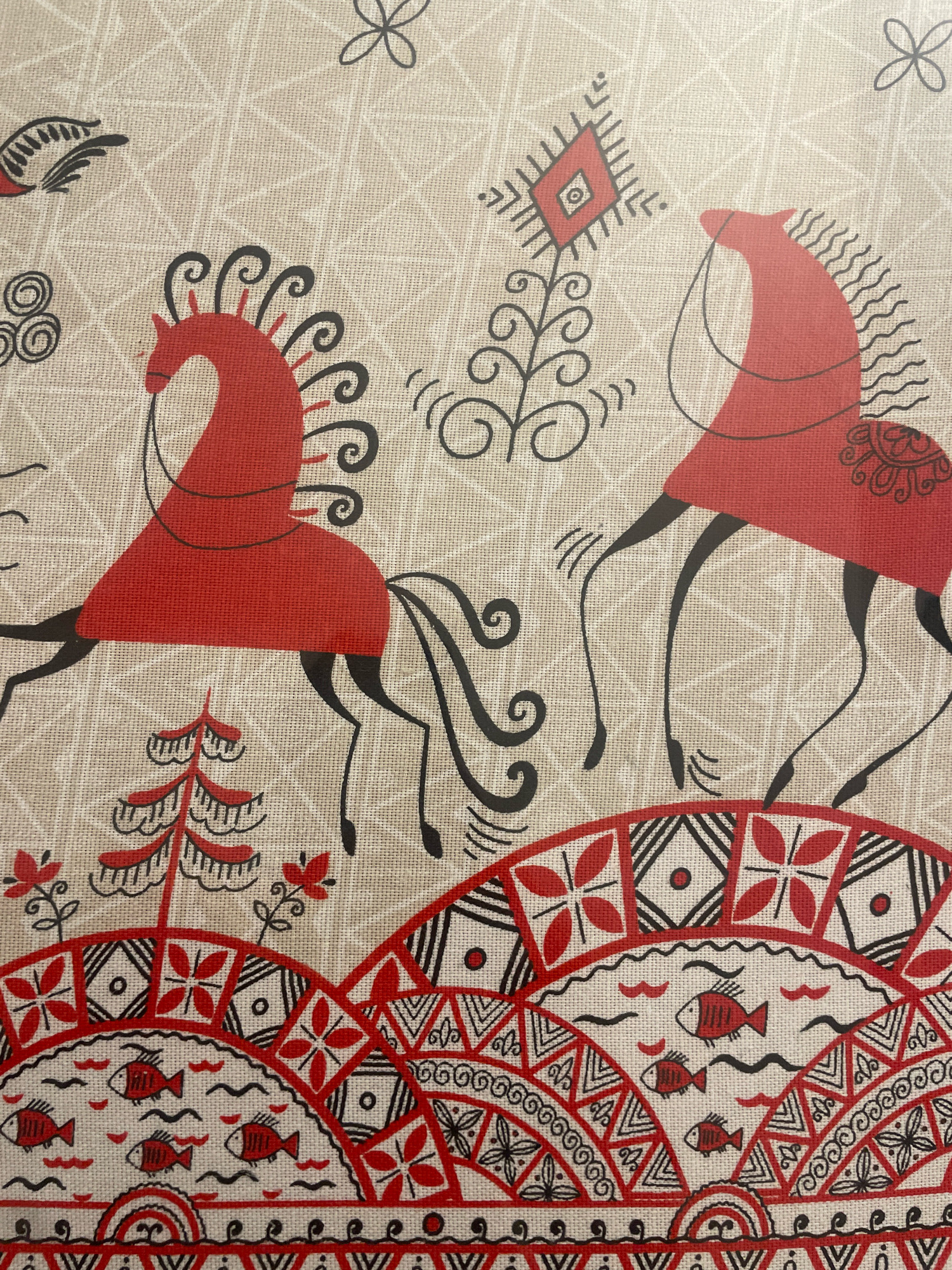 The image shows a close-up of a textile with a traditional folk art design. The design features two stylized red horses with black manes, tails, and legs. One horse is on the left, facing right, with decorative swirls emanating from its mane and a dotted pattern on its flank. The other horse is on the right, facing left, with wavy lines for its mane and a similar dotted pattern on its flank. Both horses are depicted against a beige background with a subtle grid pattern.
Below the horses, the design transitions to a landscape with stylized trees and a decorative border. The border is predominantly red and black, featuring geometric patterns, floral motifs, and depictions of fish swimming in wavy lines. The overall style is reminiscent of traditional embroidery or block printing.
There is no indication of the city of Atarfe, Spain, in the image or its context. The image is a detail of a textile and does not depict any recognizable landmarks or features associated with a specific geographical location.
