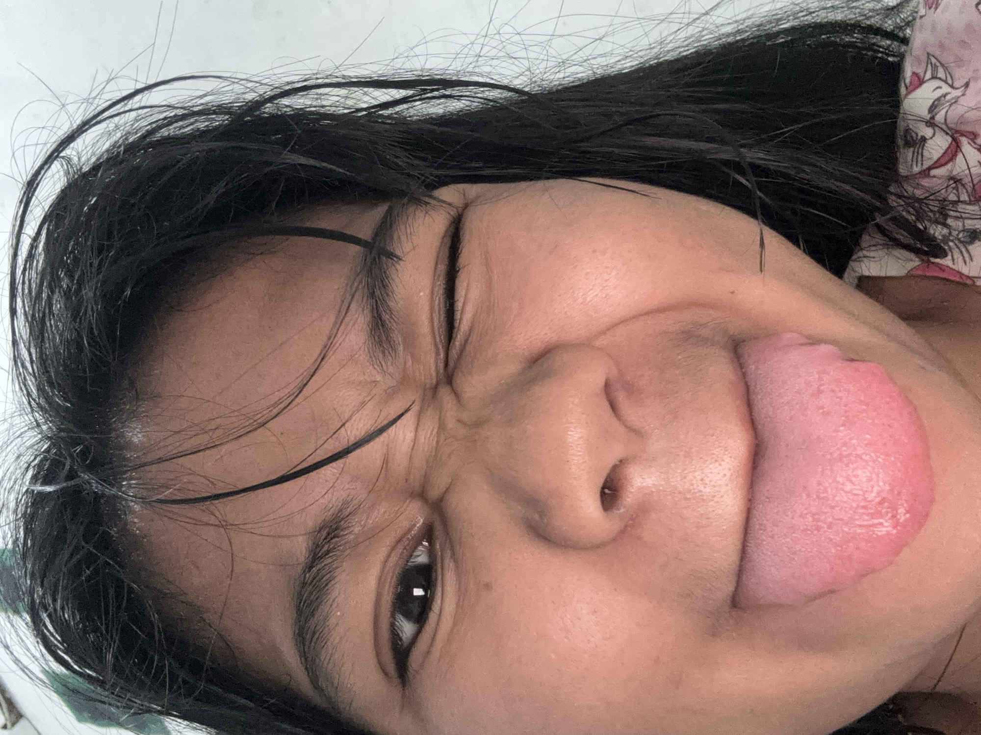 A close-up, low-angle selfie of a young woman with dark hair sticking to her face and shoulders. She is winking with her left eye and sticking out her tongue. Her skin appears slightly reddish, and there are fine hairs visible on her face and neck. She is wearing a pink and white patterned garment. The background is a plain, light-colored wall. The image was taken indoors in North Jakarta, Indonesia.