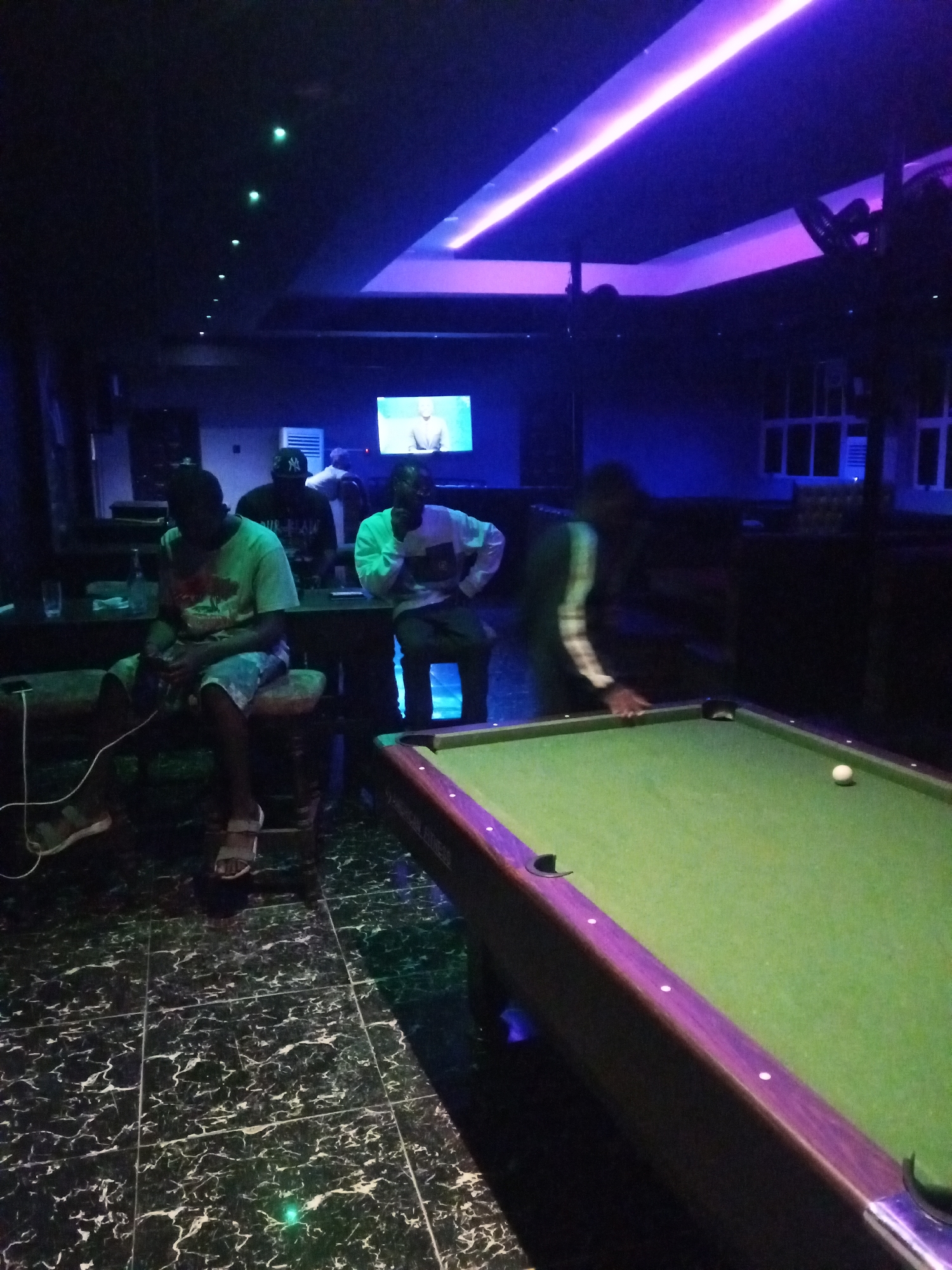 The image depicts an indoor scene, likely a dimly lit bar or entertainment venue, with a pool table in the foreground. The overall ambiance is set by colored lighting, with a prominent purple and blue glow from overhead fixtures and accent lighting on the pool table.
In the foreground, a green pool table with a wooden and purple frame is visible. A single white cue ball rests on the table. To the right, a person's hand is poised over the table, suggesting they are about to play pool.
In the mid-ground, several individuals are seated on stools or chairs around a table. One person in a light-colored t-shirt and shorts is seated on the left, with their legs crossed and a wire extending from their lap. To their right, another person wearing a dark t-shirt and a cap is seated. Further to the right, a person in a light-colored long-sleeved shirt is also seated.
In the background, a television screen displays an image of a person in a suit. The room has a dark ceiling with several small green lights. The walls appear to be dark, with some windows or panels visible on the right side. The floor is tiled with a dark, marbled pattern.
The scene suggests a casual gathering of people engaged in leisure activities. The location is identified as Lokoja, Nigeria.