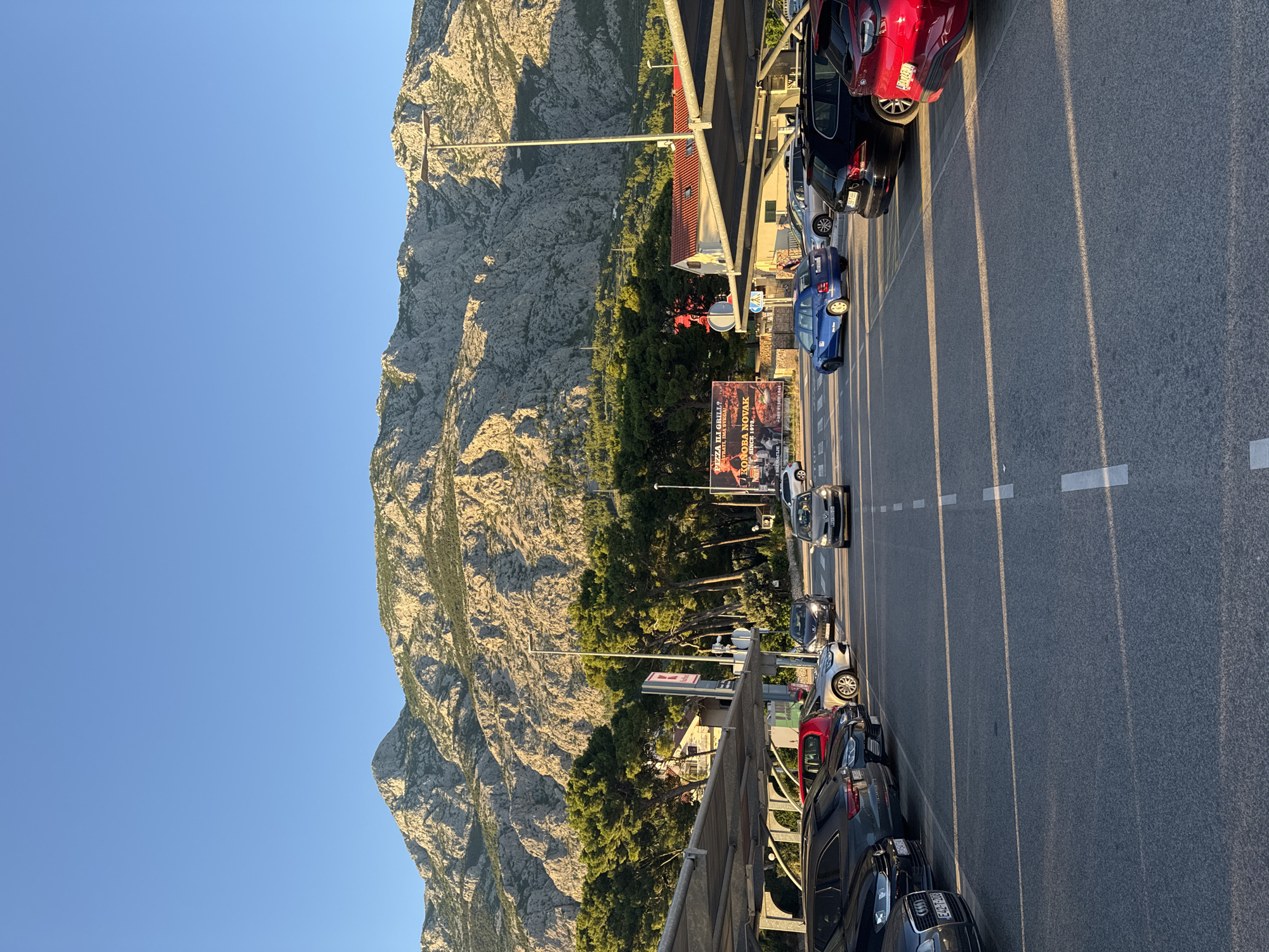 The image depicts a view of a parking lot in Makarska, Croatia, with a large, rugged mountain range as the backdrop. The sun is low in the sky, casting long shadows across the asphalt. Several cars are parked under a covered structure on the left side of the frame, and more cars are visible driving or parked in the open area. A large billboard advertising "PIZZA U GRILL?" and "KONOBA NOVAK" is prominent in the middle ground, with a red-tiled roof building and green trees surrounding it. The mountain range is textured and rocky, with patches of vegetation. The sky is clear and blue, indicating a pleasant day.