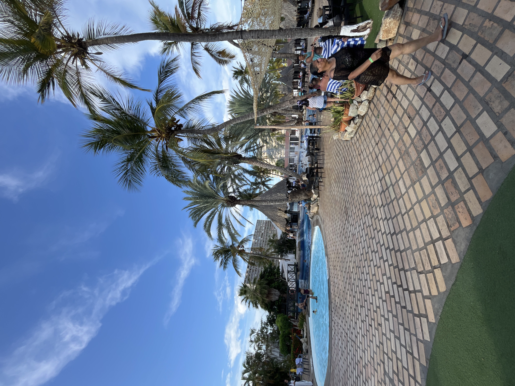 The scene depicts a tropical resort area in Porlamar, Venezuela. A kidney-shaped swimming pool is the focal point, surrounded by a brick-paved patio. Palm trees line the pool area, casting shadows and contributing to the tropical atmosphere. Several people are present: some are swimming in the pool, while others are lounging on chairs near the pool or sitting at tables under thatched umbrellas. A woman in a black lace cover-up is relaxing on a chair. In the background, a multi-story building is visible. The sky is blue with scattered clouds. The environment suggests a relaxed vacation setting.