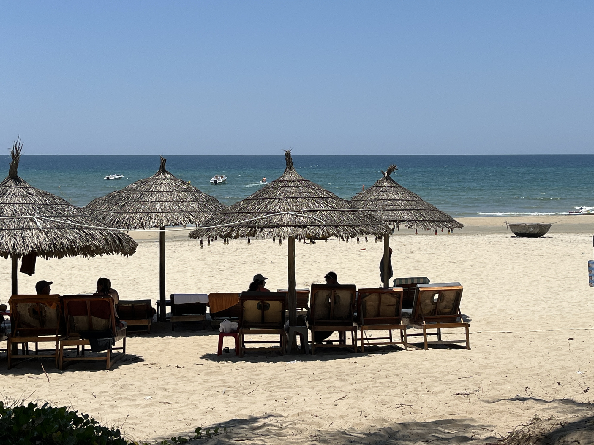 The image depicts a beach scene in Đà Nẵng, Vietnam. Four thatched umbrellas provide shade for beachgoers in wooden chairs. People are seated under the umbrellas. The beach is sandy, leading to a calm ocean with several boats in the distance. A round boat sits on the beach to the right.