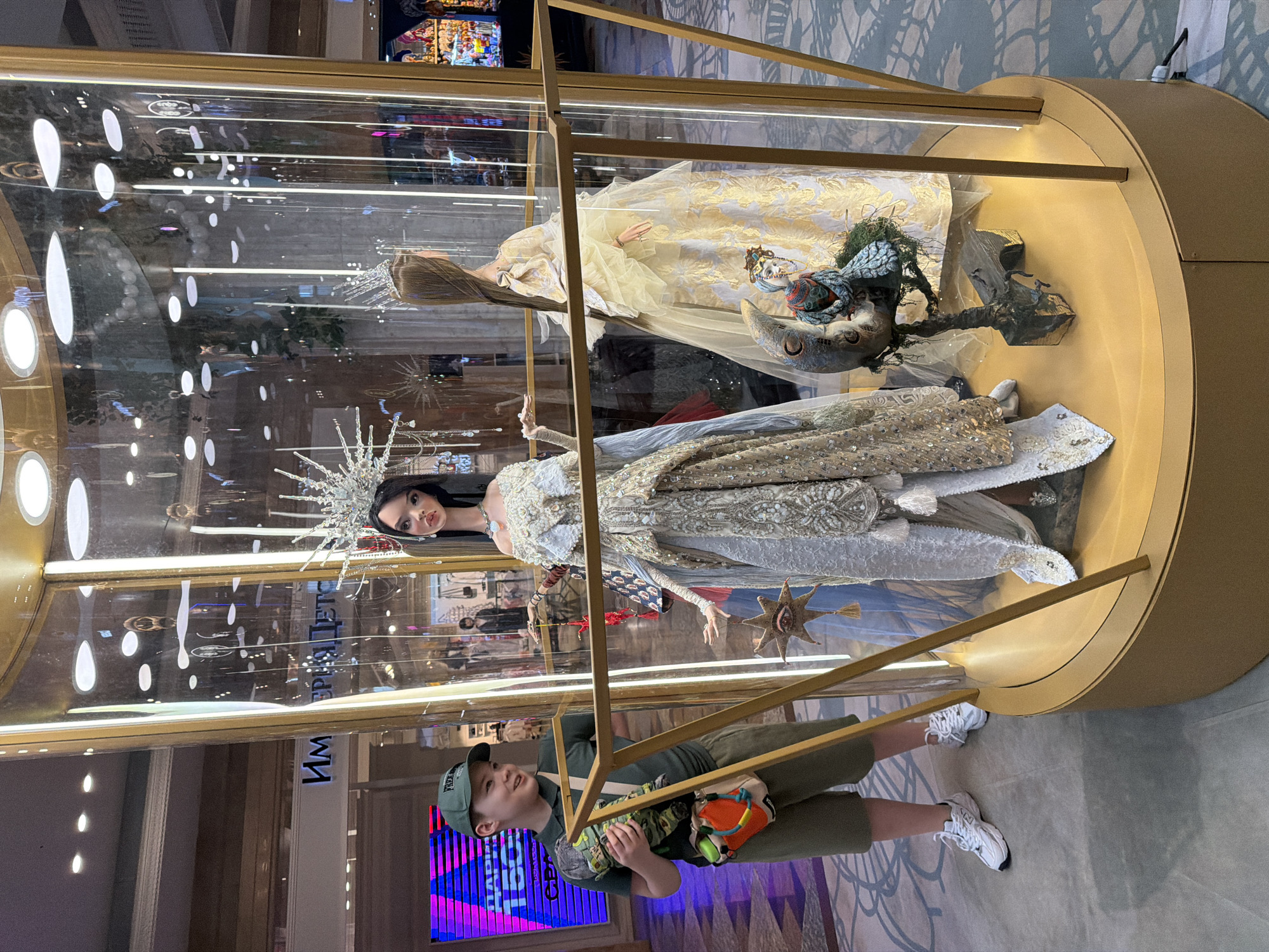 A boy stands in front of a glass display case containing two mannequins wearing elaborate dresses, located in a Moscow, Russia shopping mall. The boy is wearing a green t-shirt, green cap, green shorts, and white sneakers. The lower mannequin wears a silver dress with a spiky crown, while the upper mannequin wears a gold dress. The display case has a gold base and a light strip on the top edge. A store sign is visible in the background.