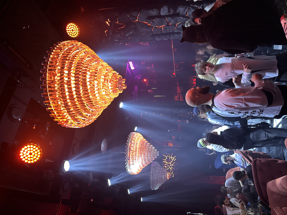 The image portrays an active indoor nightlife venue identified as YATOO Bar (ЯТОО) in Moscow, Russia. The environment is dimly lit with elaborate theatrical lighting, featuring three multi-tiered crystal chandeliers emitting amber light, multiple red LED spotlights mounted on the ceiling, and several white spotlights projecting visible beams through a smoky or hazy atmosphere.
Numerous individuals, primarily young adults, occupy the space. In the midground-right, a bald male with a beard, wearing a light-colored long-sleeved shirt, faces left while engaged in interaction with a younger blonde-haired male. The blonde male wears a white and black long-sleeved athletic-style shirt with "CASHER" text. To his right, an individual is observed from behind, wearing a black top and a headpiece with ear-like protrusions. Further left, a person in a striped beanie stands near a female with dark hair, dressed in a dark top and light-colored jeans. On the far left, several patrons are seated on plush red armchairs around small tables.
The background reveals additional individuals standing and socializing. A wall on the right side features dark, textured, rock-like panels with red light emanating from crevices. An elevated area, possibly a DJ booth, is visible in the deep background, displaying a neon sign with the Cyrillic text "ЯТОО."