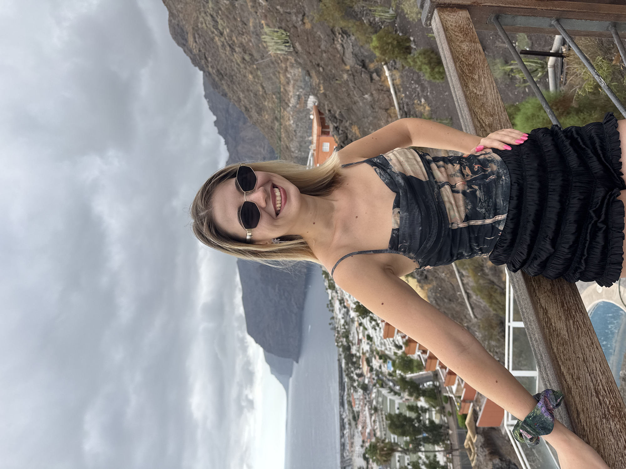 A woman with blonde hair and sunglasses stands on a wooden railing overlooking a coastal town with a harbor and dramatic cliffs in the background. The sky is overcast. She is wearing a patterned top and black ruffled shorts. Her right hand is on her hip, and her left arm is resting on the railing.
