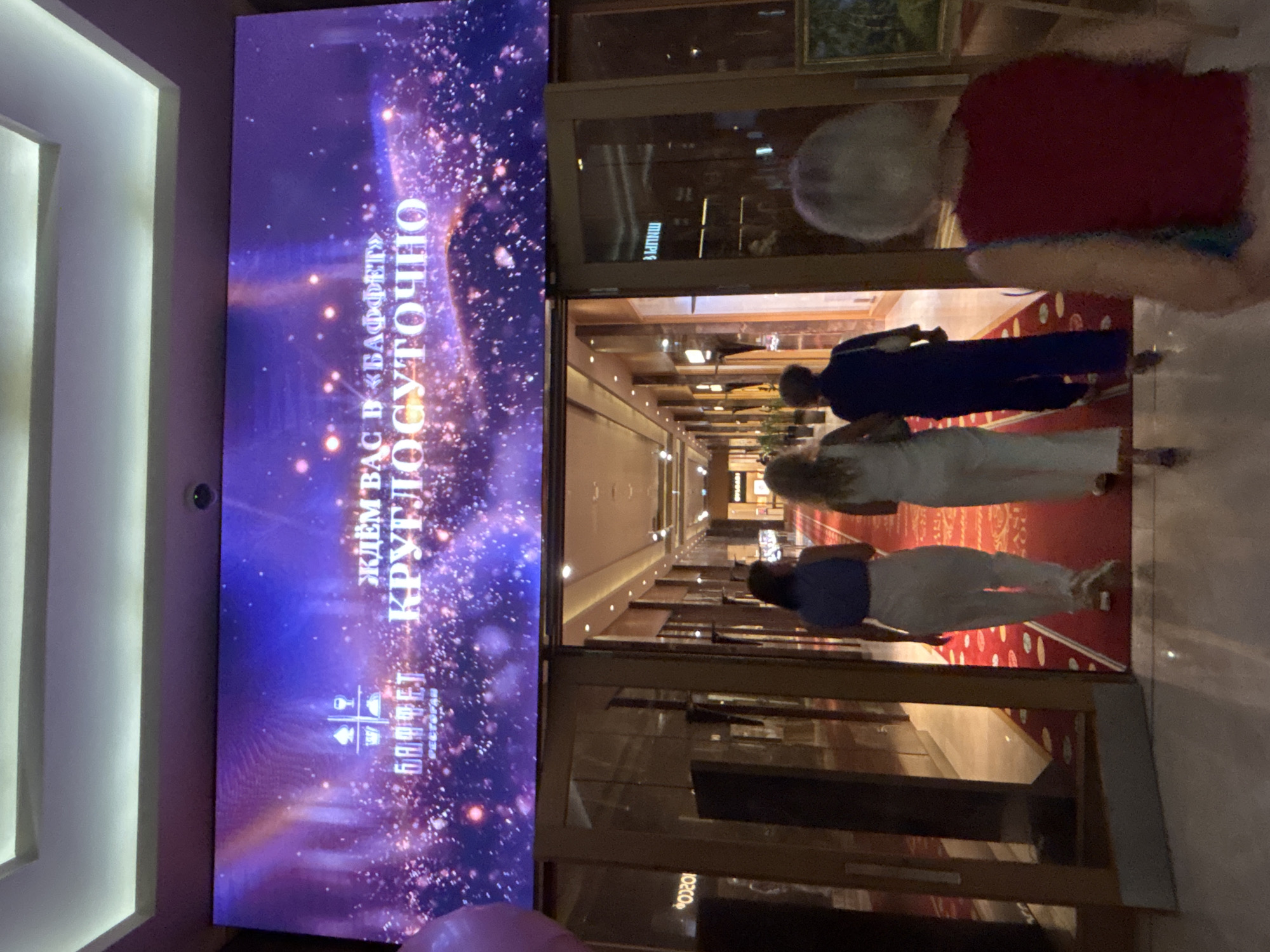 The image captures a scene inside a building, likely a hotel or entertainment complex, in Krasnaya Polyana, Russia. Dominating the upper portion of the image is a large LED screen displaying vibrant purple and pink abstract patterns with glittering lights. Text on the screen is in Russian, indicating "ЖДЁМ ВАС В «БАФФЕТ» КРУГЛОСУТОЧНО" which translates to "WE ARE WAITING FOR YOU AT 'BUFFET' 24/7". Below the screen, a wide entrance with glass doors leads into a brightly lit corridor. A red carpet runs down the center of the corridor, with decorative circles along its edges.
Three women are visible walking into the building, away from the camera, down the red carpet. The woman in the foreground on the left wears a blue top and white pants. The middle woman wears a white jumpsuit. The woman on the far right, also walking away from the camera, has grey hair and wears a red top and appears to be holding a blue bag. The environment suggests a luxurious establishment, possibly a casino or a high-end restaurant, given the prominent branding and the upscale interior design with polished floors and decorative lighting. The presence of shops or display cases can be seen through the glass doors on the left. The overall atmosphere is one of welcoming guests to a round-the-clock establishment.