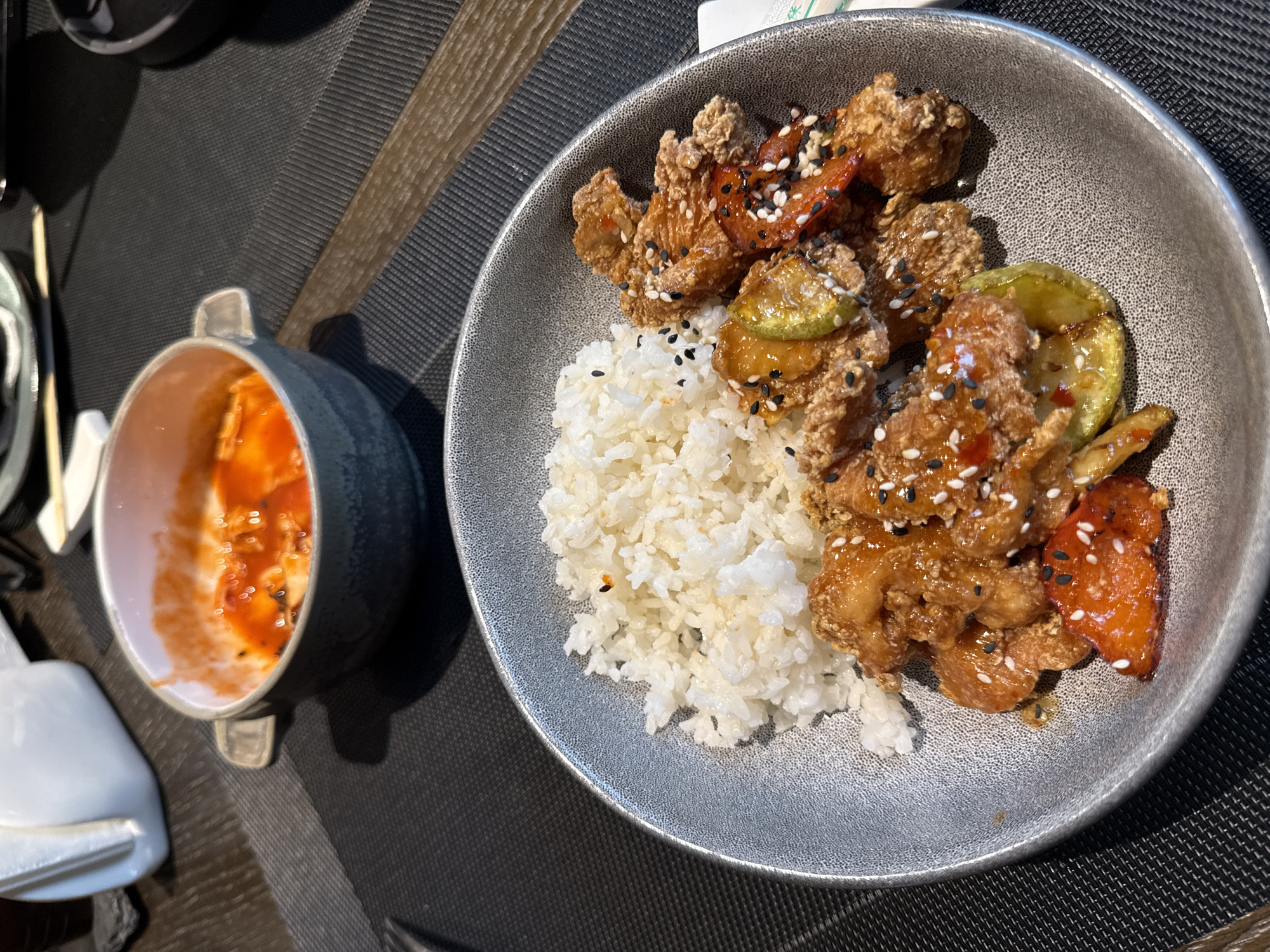 This media file captures a close-up, elevated view of a prepared meal served on a dark grey, textured placemat, likely in an indoor restaurant or cafe setting in Moscow, Russia.
The central subject is a large, dark grey/silver bowl filled with food. The bowl contains a generous portion of white, fluffy rice on one side. On the other side are several pieces of crispy, deep-fried food, likely chicken or a similar protein, coated in a shiny, light brown glaze and generously sprinkled with both white and black sesame seeds. Mixed in with the fried pieces are stir-fried or grilled vegetables, specifically bright green slices resembling zucchini and reddish-orange pieces that appear to be bell peppers, also coated in the glaze and sesame seeds.
To the left, a smaller, dark grey bowl with a light pink interior holds a reddish-orange, somewhat chunky sauce, possibly a spicy dipping sauce or kimchi, also garnished with a few black sesame seeds. Adjacent to this smaller bowl, a light-colored object, possibly a utensil rest or part of a serving set, is partially visible. Near the main bowl, a piece of white paper or a coaster shows blurry green text, with one discernible character appearing to be "米," which means "rice" in East Asian languages.
The lighting is consistent with indoor artificial light, casting soft shadows. The food appears fresh and neatly arranged, indicating a meal recently served. No people are visible in the image.