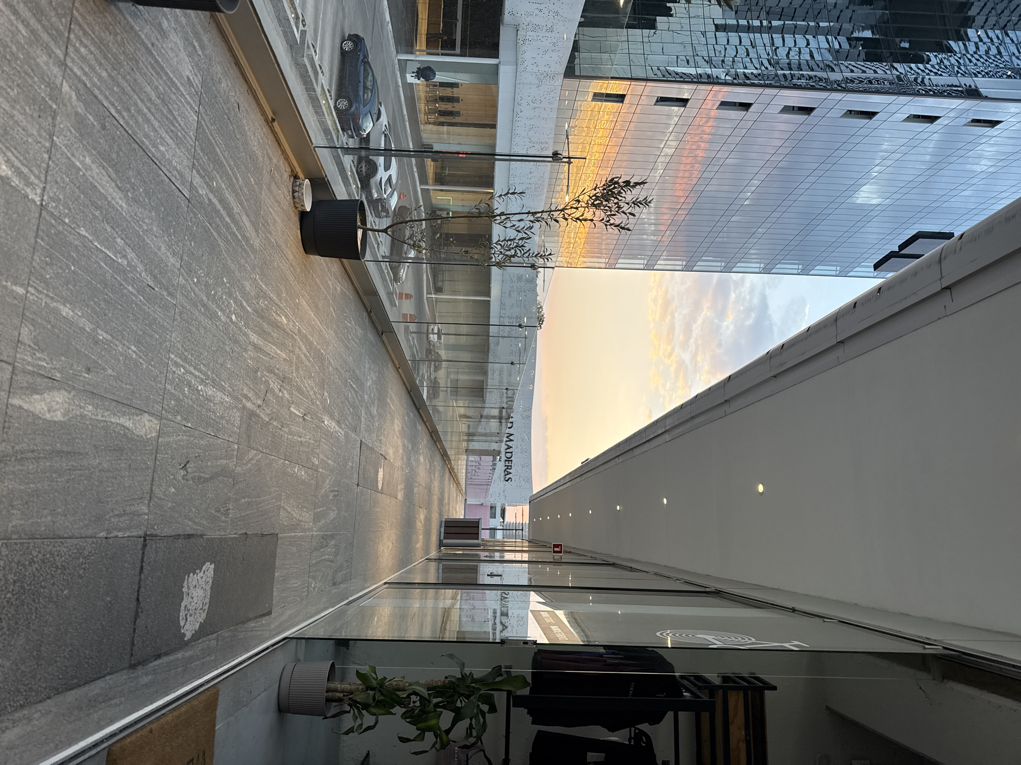 The image presents an elevated outdoor pedestrian promenade within a modern commercial or mixed-use development in Puebla, Mexico, during late afternoon or early evening. No individuals are present.
On the left, a tall, multi-story glass-clad skyscraper reflects the sky, displaying prominent orange, pink, and blue hues of the sunset. Below this structure, a parking area contains at least four parked vehicles: a dark blue hatchback, a white SUV, a silver-grey SUV, and a dark SUV. Orange traffic cones are partially visible on the ground level. A white concrete facade with a perforated pattern borders the parking area beneath the skyscraper. A single, slender potted tree with dark foliage is situated on the promenade, positioned in front of a clear glass railing that extends along the edge of the walkway.
The promenade surface consists of large, light grey rectangular paving tiles with a visible striated texture. Recessed circular lights are embedded in the white ceiling extending above the promenade. The right side of the walkway is lined with several glass storefronts. Reflections in these windows show the opposite side of the promenade. Inside one visible store, dark clothing items hang on racks, and a potted plant in a light grey container is present. A white circular logo is affixed to one of the store windows. A small, decorative, patterned cylindrical object lies on the tiled floor near the glass railing.
In the background, across an open area beyond the parking, a building with a white facade prominently displays the name "CIUDAD MADERAS." Further back, another light-colored building with a pinkish rooftop section is visible beneath a sky with scattered clouds illuminated by the low sun.