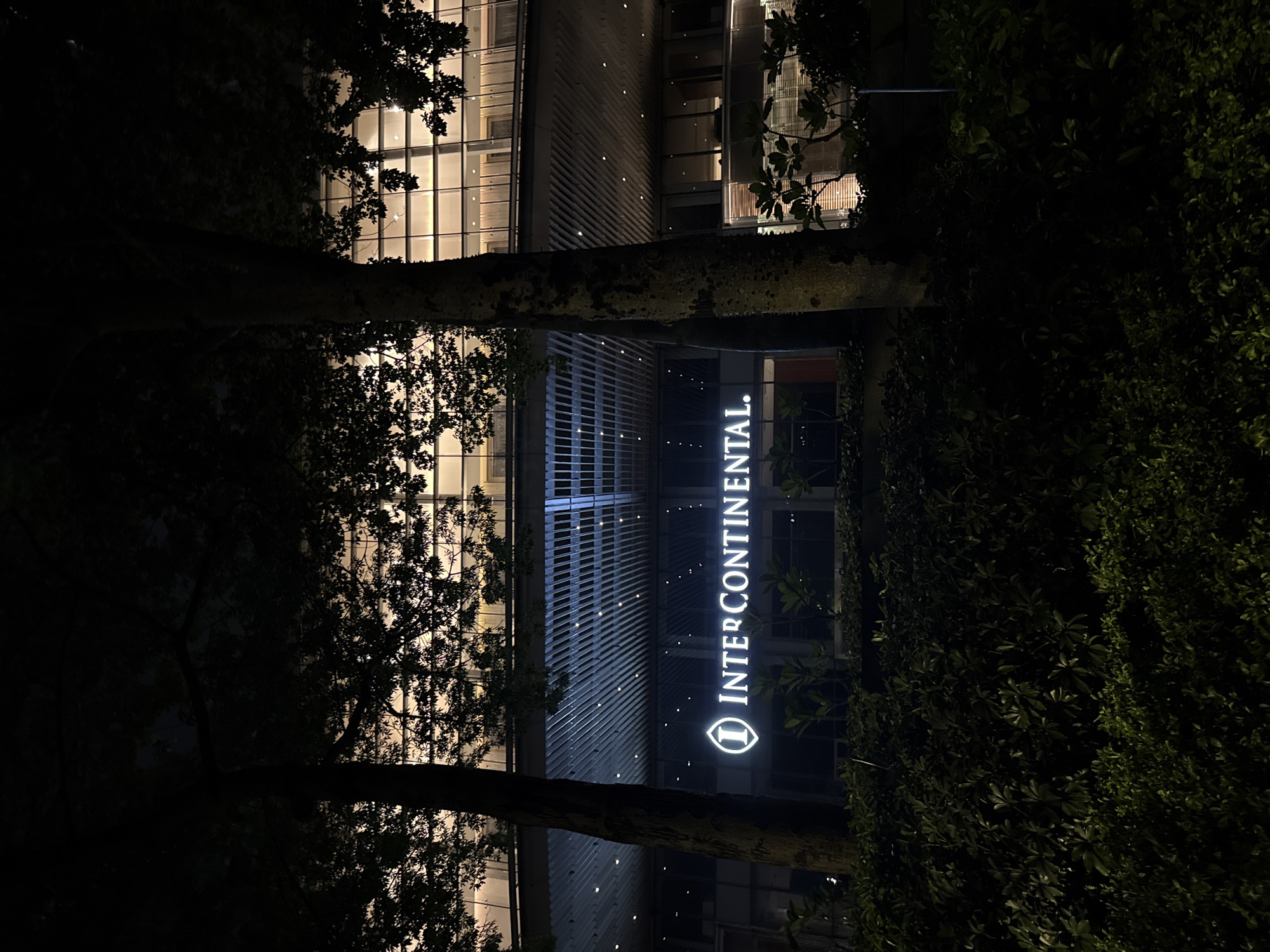 The image shows the exterior of the InterContinental hotel at night in Haizhu District, China. The hotel building features a modern design with a facade of windows and metallic louvers, illuminated from within. A prominent, glowing sign displays the "InterContinental" logo, with the distinctive "i" symbol. The foreground is dominated by dark silhouettes of trees and lush green foliage, suggesting a garden or park area surrounding the hotel. The time of day is clearly night, with artificial lighting from the building and the sign being the primary sources of illumination. The scene evokes a sense of tranquility and sophistication, typical of a high-end hotel setting.