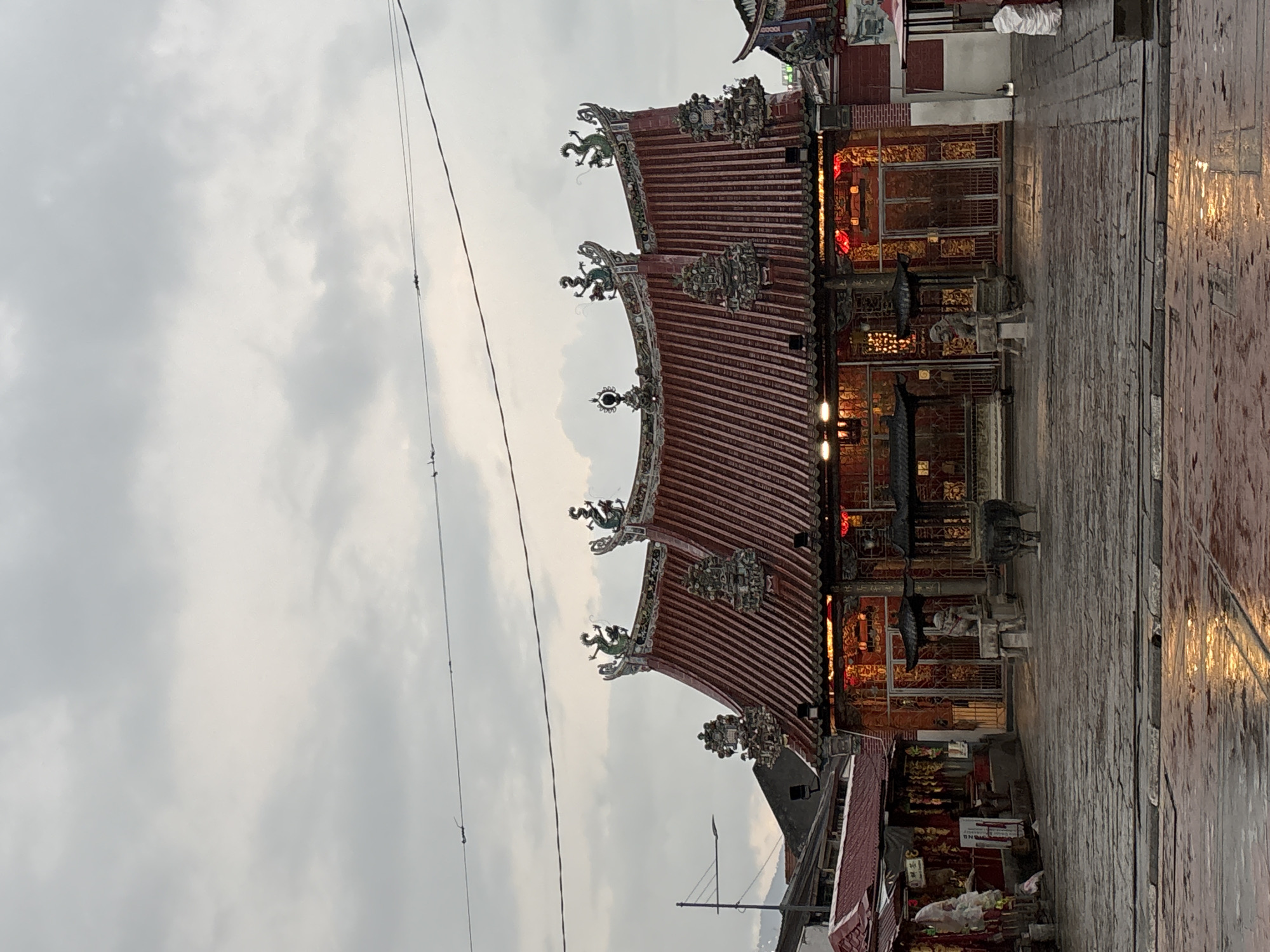 The image shows the exterior of a traditional Chinese temple, likely located in George Town, Malaysia, given its architectural style. The temple features a prominent red, multi-tiered roof adorned with ornate dragon sculptures and decorative elements. The facade is decorated with red lanterns and intricate golden carvings. In front of the temple, there is a paved courtyard that appears wet, suggesting recent rain, with reflections of lights visible on the surface. Two stone lion statues flank the entrance, and a large incense burner stands in the foreground. To the left of the main temple structure, there are smaller buildings with red roofs and displays of various items, possibly for sale or offerings. The sky above is overcast with grey clouds. No people are visible in the scene.
