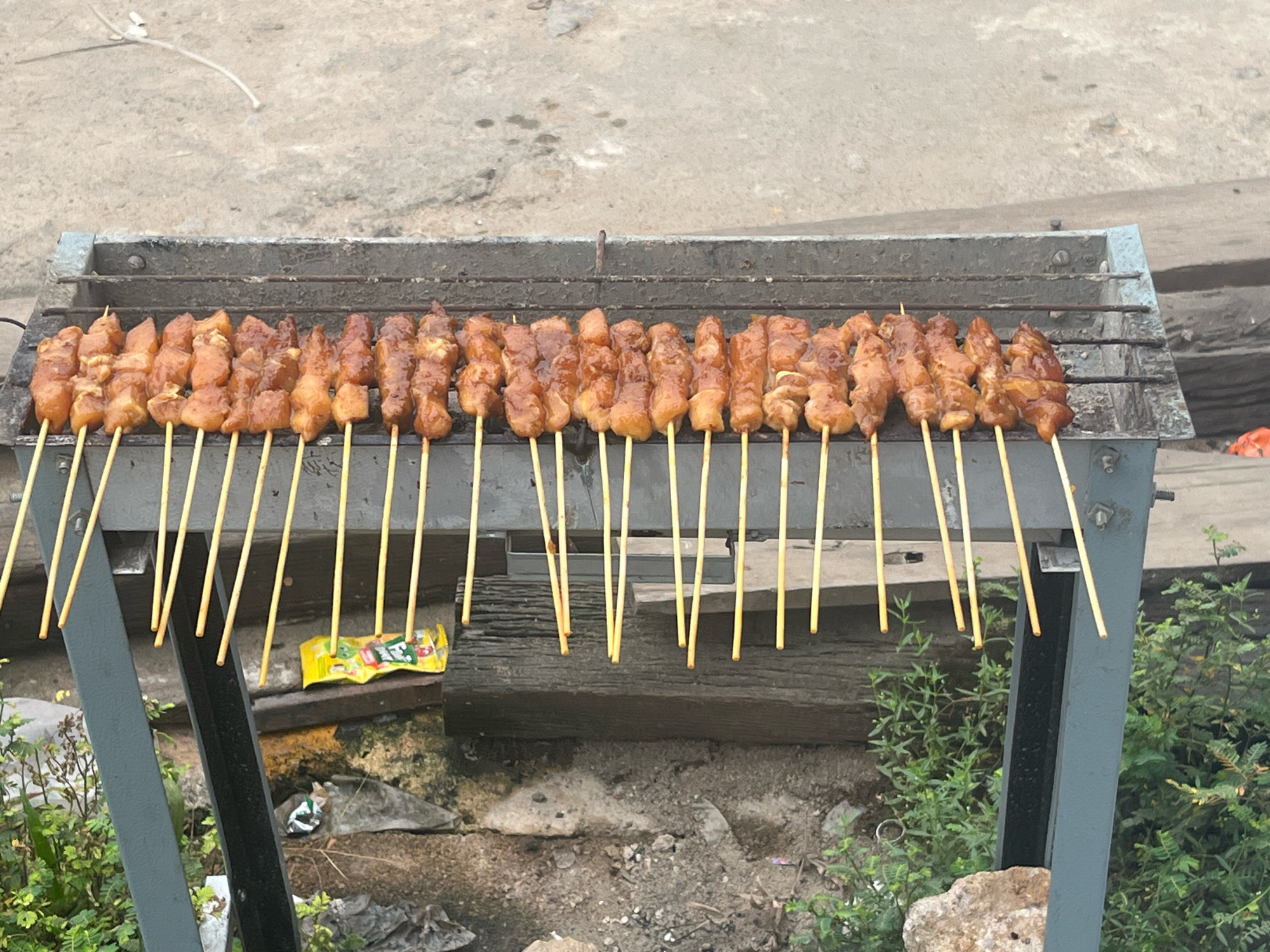 The image shows a close-up view of a charcoal grill laden with rows of uncooked meat skewers, likely satay, in Meliau Hilir, Indonesia. The skewers, each with pieces of marinated meat, are neatly arranged on the metal grill. The grill itself appears to be a portable, metal structure with visible rust and char marks, indicating its use. In the background, a concrete surface is visible, possibly a street or a courtyard. The environment appears to be outdoors, with some greenery and debris scattered around the base of the grill. A yellow and red candy wrapper lies on the ground near the grill. The overall scene depicts a street food vendor preparing to cook.