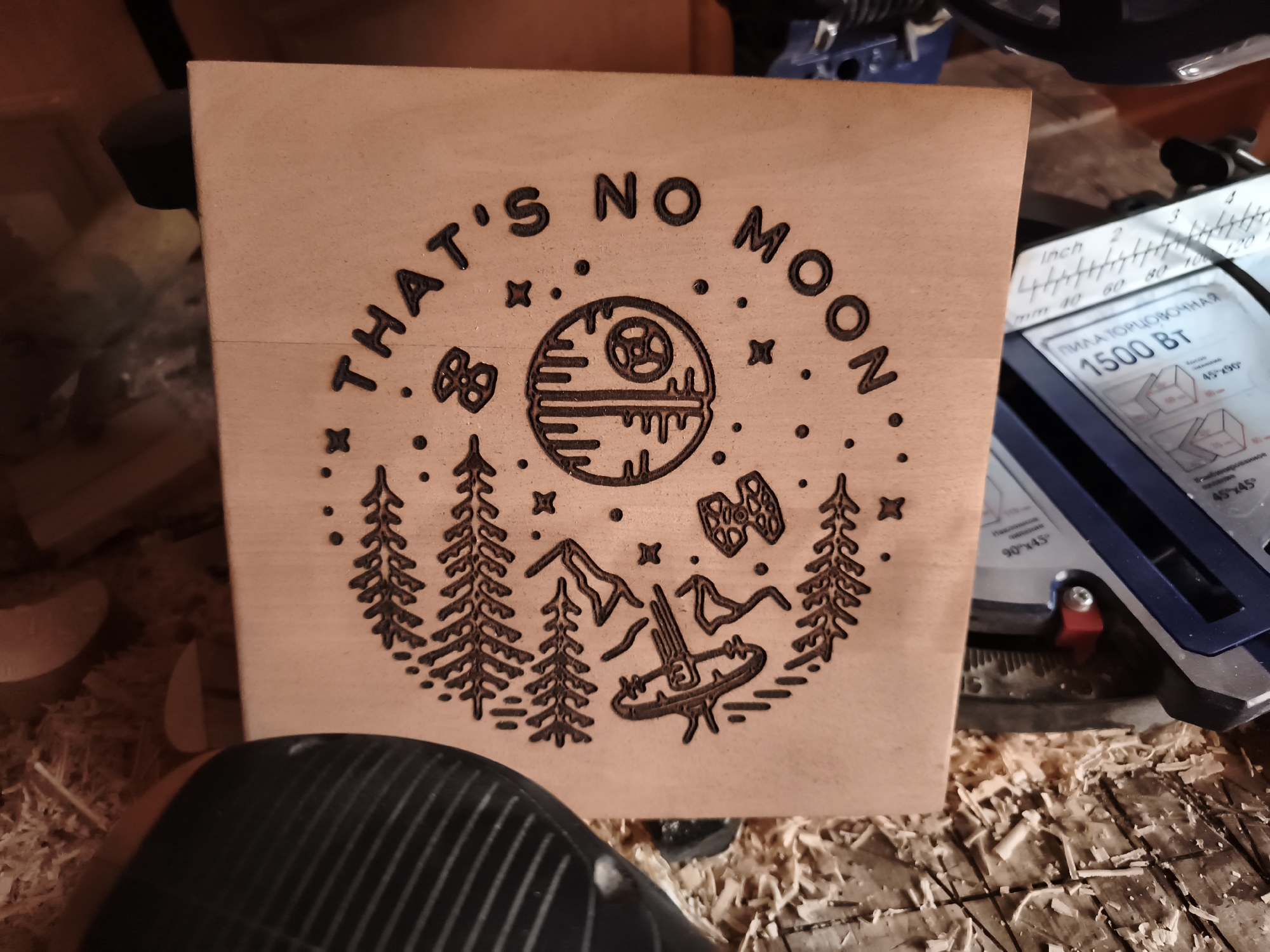 A close-up, overhead shot captures a wooden box with a pyrography design. The design features the phrase "THAT'S NO MOON" in an arc around a depiction of the Death Star, with smaller Star Wars icons like a TIE fighter and X-wing. The scene is set amidst wood shavings and tools, suggesting a workshop environment. A portion of a miter saw with a visible measuring scale is visible on the right side of the frame. The lighting is dim and appears to originate from overhead. The location is stated to be in Гаранскі сельскі Савет, Belarus.