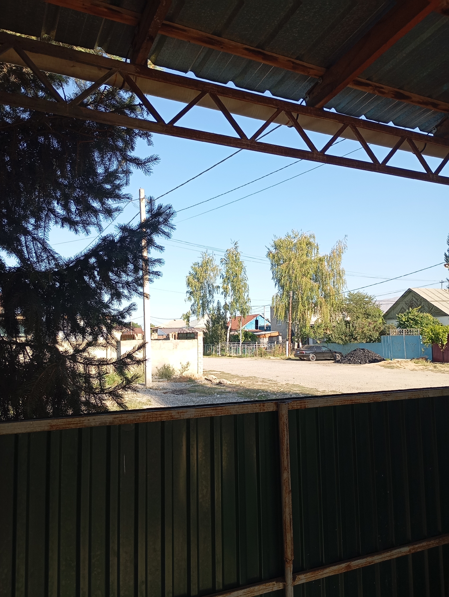 The image depicts a view through a covered porch or shelter with a corrugated metal roof and a wooden support structure. In the foreground, the dark green, corrugated metal of a fence is visible, partially obscuring the scene. To the left, a large, dark green evergreen tree branches into the frame. Beyond the fence and tree, a street or courtyard area is visible. A light-colored concrete wall stands to the left of this area, with a utility pole nearby.
Further into the scene, a dark-colored sedan is parked on a gravelly surface. To the right of the car, a large pile of dark, lumpy material, possibly coal or waste, is present. Several trees with light green foliage are scattered throughout the area, and residential buildings with various rooflines and colors are visible in the background. A light blue metal gate with decorative elements is also present. The sky is clear and blue. The overall impression is a residential street scene in a rural or semi-rural area. The location is Talas City, Kyrgyzstan.
