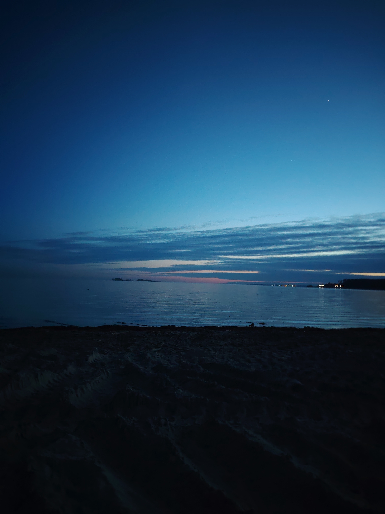 The image depicts a coastal scene at dusk. The sky transitions from a deep blue overhead to a lighter blue near the horizon, where thin, striated clouds are present. A small, bright object, possibly a star or planet, is visible in the upper right quadrant of the sky. Below the sky, a body of water stretches across the middle of the image. The water is calm, with faint reflections of the sky and clouds. In the distance, there are small landmasses or islands, and further along the shoreline, some lights are visible, suggesting a town or settlement. The foreground is a dark, textured surface, likely sand, with visible tracks or patterns. The overall scene appears to be a tranquil and remote location, possibly a beach. The location is in the city of Unknown, Unknown.