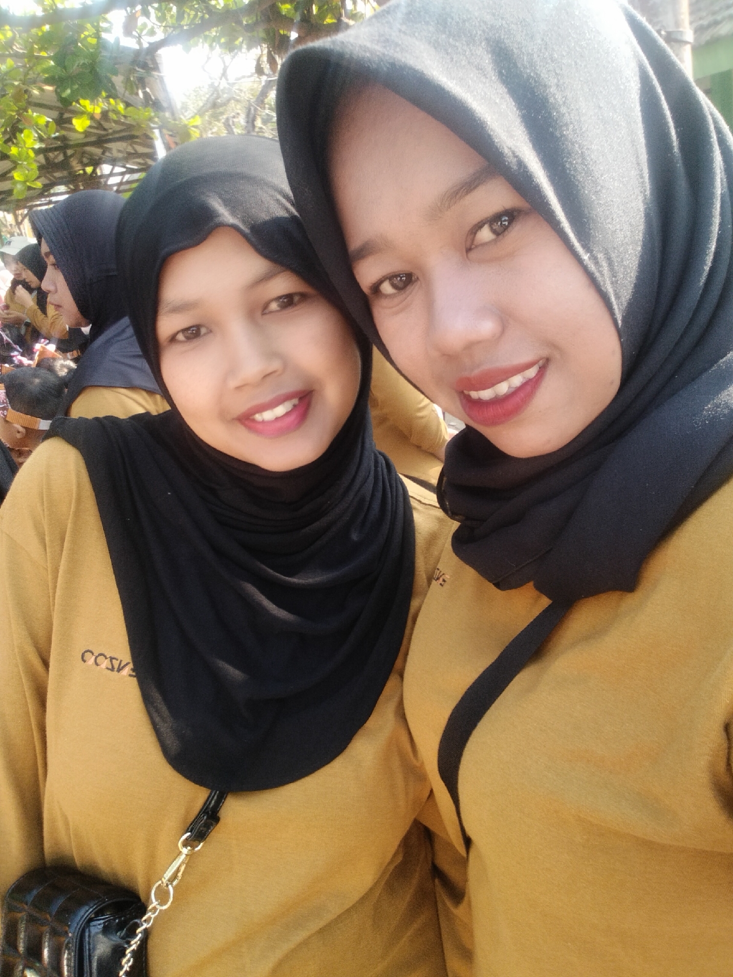 In a medium shot, two young women wearing black hijabs and mustard-colored shirts are posing for a selfie outdoors. The woman in the foreground is on the right, with her face angled towards the camera and her mouth slightly open in a smile, revealing her teeth. She has light brown eyes and is wearing red lipstick. The woman on the left is also looking at the camera, her head tilted slightly to the right. She has a wide smile with her teeth visible. Both women have dark eyebrows.
Their shirts have an inscription in black letters on the left chest area, which appears to read "CCSVE" or similar. The woman on the left has a black quilted purse with a gold chain strap hanging from her shoulder, with the strap attached to a gold clasp visible on her shirt. The background is blurred but suggests an outdoor setting with greenery and possibly other people further in the distance. Sunlight is casting bright highlights, particularly on the faces and clothing of the subjects, indicating it is a sunny day. The image appears to be taken in Puspo, Indonesia, given the provided context.