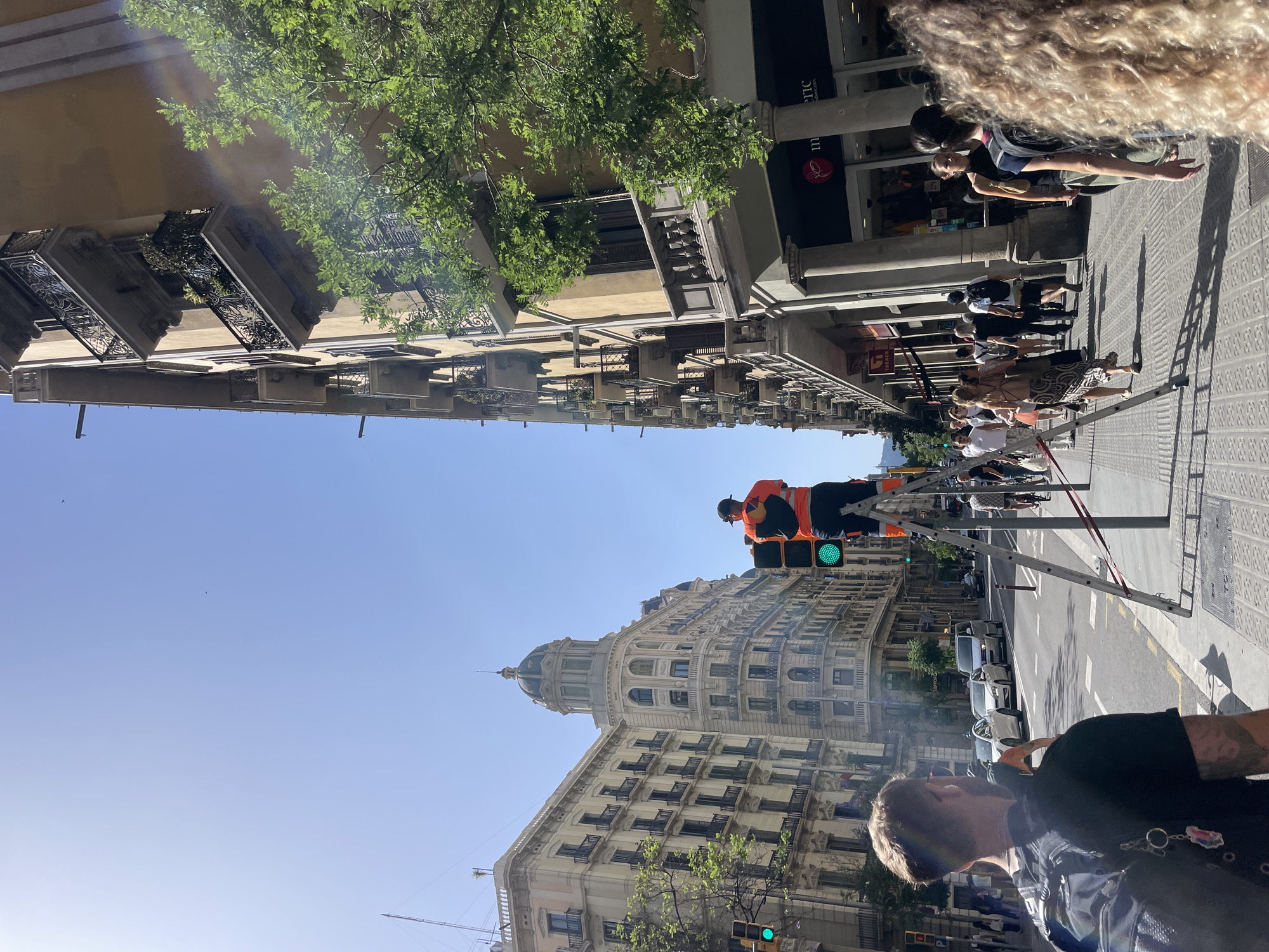 This media file depicts a sunny day in Barcelona, Spain, as suggested by the clear blue sky, bright lighting, and shadows cast on the ground. The scene is an urban street with a mix of architectural styles. In the foreground, several people are visible, some walking and others standing. One person in an orange vest is standing near a ladder, possibly involved in some sort of work or setup. In the mid-ground, traffic, including cars and what appears to be a small truck, is moving along the street.
The buildings lining the street are ornate, with many windows, balconies, and decorative elements, suggesting an older European city. One prominent building features a dome and multiple stories of arched windows. Another building has a repeating pattern of balconies with dark railings. Lush green trees provide shade in some areas.
The image captures a bustling street scene with people engaged in various activities, creating a sense of everyday life. The bright sunlight indicates it is likely midday. There is no explicit text visible in a legible manner within the image. The overall mood appears to be one of activity and a typical day in a vibrant city.