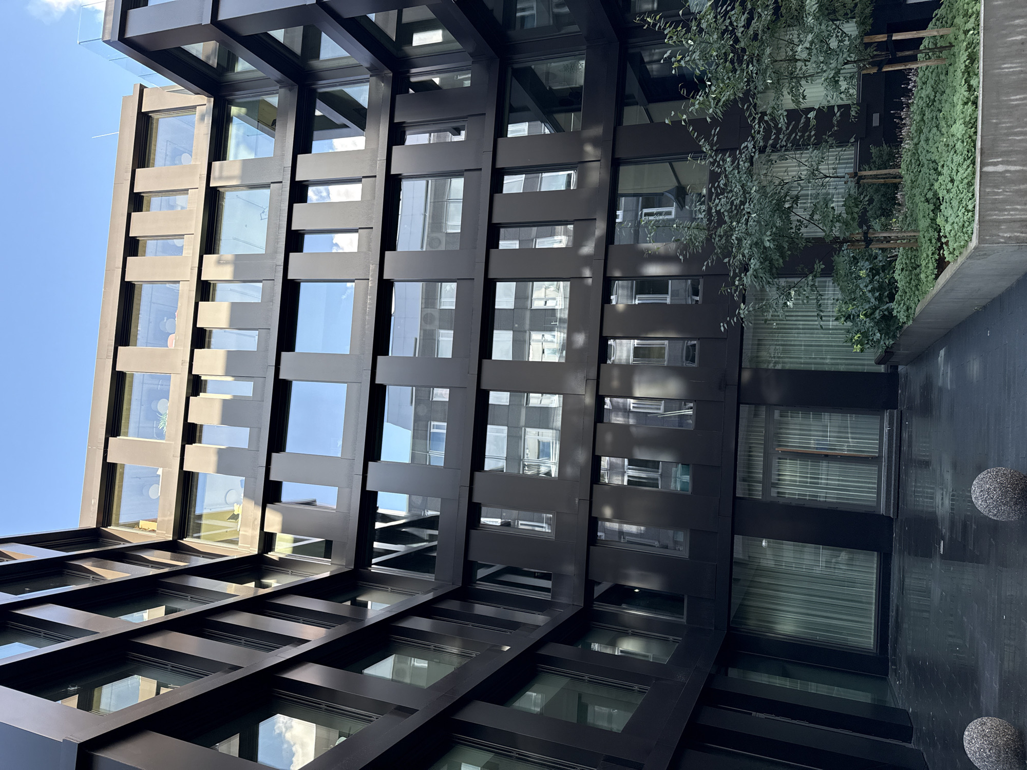 The image depicts the modern architectural design of the Rotermann Quarter's shopping center in Tallinn, Estonia. The building features a dark facade with numerous large, rectangular windows, many of which reflect the blue sky and clouds. The perspective is looking upwards from a courtyard area paved with dark, wet tiles, suggesting recent rain. Two spherical, grey, pebble-covered bollards are visible in the foreground. To the right, a raised planter with lush green ground cover and several young trees with wooden stakes stands against the building. The building's design is characterized by repeating geometric patterns created by the window frames and dark structural elements.