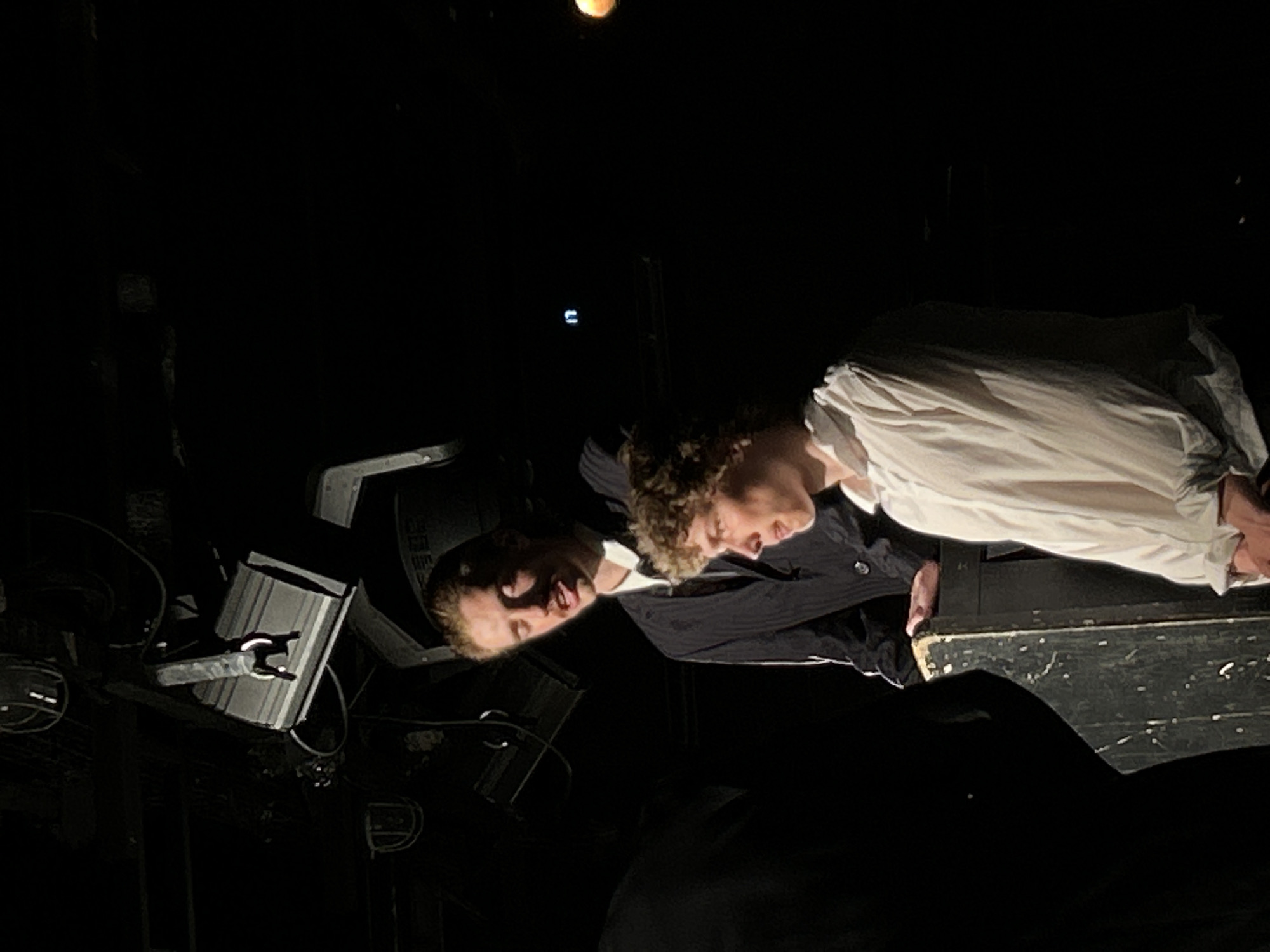 The image depicts two men, one in a white shirt and the other in a dark suit, leaning over what appears to be a surface, possibly a table or a piano. The scene is dimly lit, and there are stage lights and other equipment visible in the background, suggesting a theatrical or performance setting. The location appears to be Unknown, Unknown.