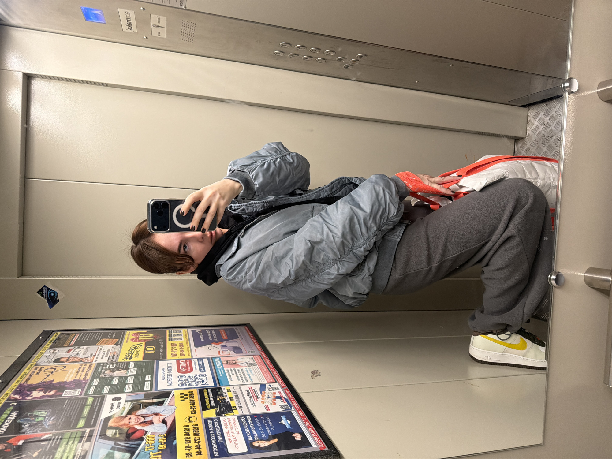 A person in an elevator takes a selfie in front of a mirror. The person is wearing a gray bomber jacket, gray sweatpants, and white sneakers with yellow accents. The elevator has various advertisements on the wall, some of which have Russian text. The OCR suggests the location might be Ust-Ilimsk, Russia, based on the Cyrillic characters and the word fragments extracted from the poster.