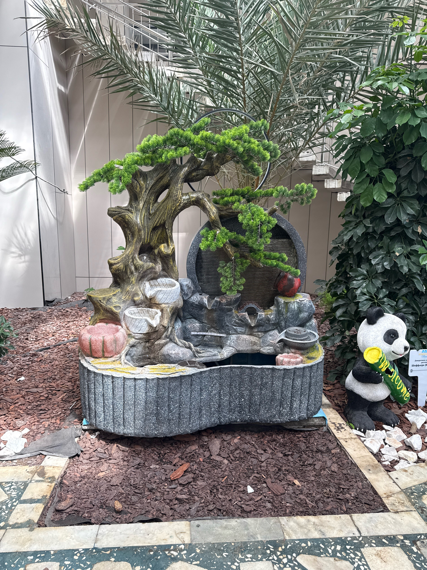 In Bosteri, Kyrgyzstan, a decorative outdoor fountain sits amidst a garden. The fountain, designed to resemble a tree with cascading water features, is made of artificial stone and concrete. A red ornamental fruit is placed on the fountain's rockery. To the right of the fountain, a ceramic panda statue holds a green sign with yellow lettering that reads "Welcome". The area is covered with dark brown mulch and bordered by tiled pathways. Lush green foliage and palm fronds surround the scene, with a building visible in the background.