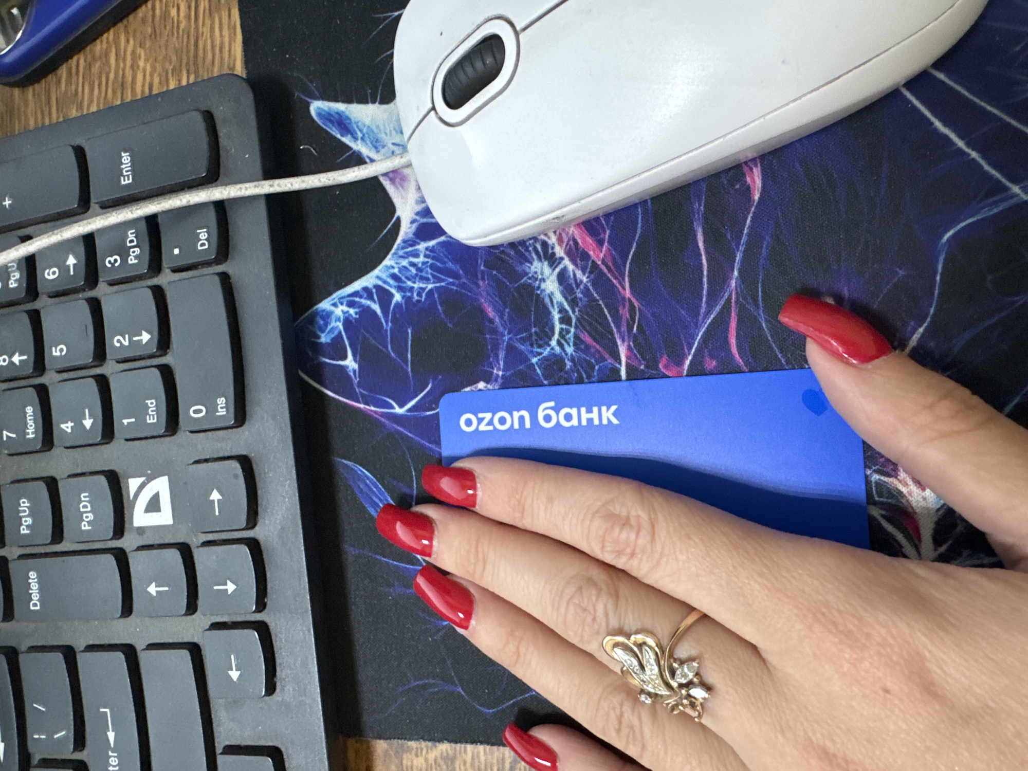 A close-up, overhead shot shows a woman's left hand with long, red-painted nails and a gold and diamond ring on her ring finger. The hand is positioned over a blue card with white text that reads "ozon 6aнк". The card rests on a mousepad with a vibrant, abstract design of blue, pink, and white lightning-like streaks. To the right of the mousepad is a white computer mouse. The background consists of a black keyboard with visible keys labeled "Enter", "Pg Dn", "Pg Up", "Delete", and various arrow keys. The scene is indoors, on a wooden surface, likely a desk.