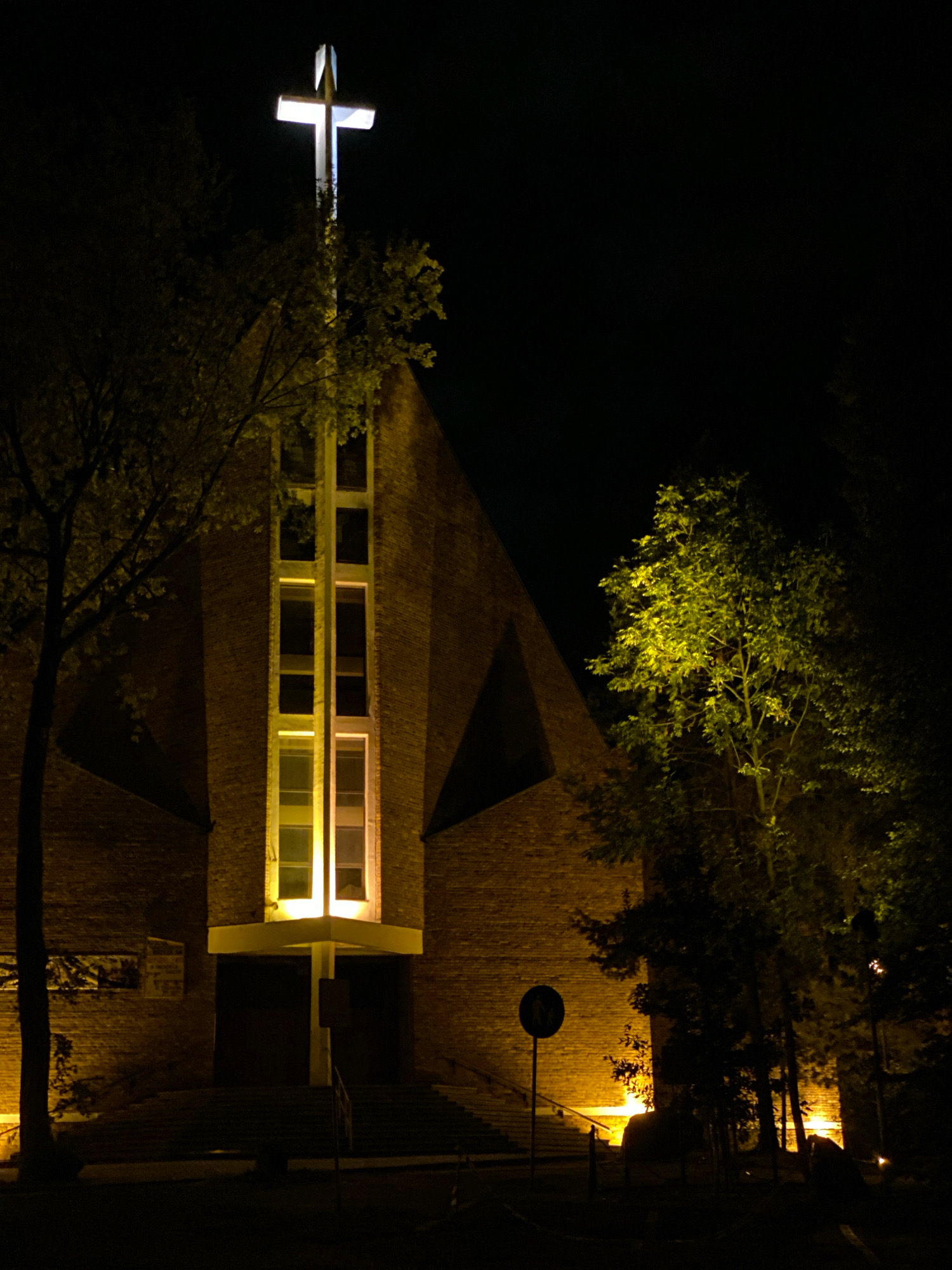 The image shows a church at night, with a illuminated cross on top of its facade. The church is made of brick and has a large set of stairs leading to its entrance. The facade is illuminated by yellow lights, and there are trees on either side of the church. The sky is dark. The church appears to be located in the city of Katowice, Poland.