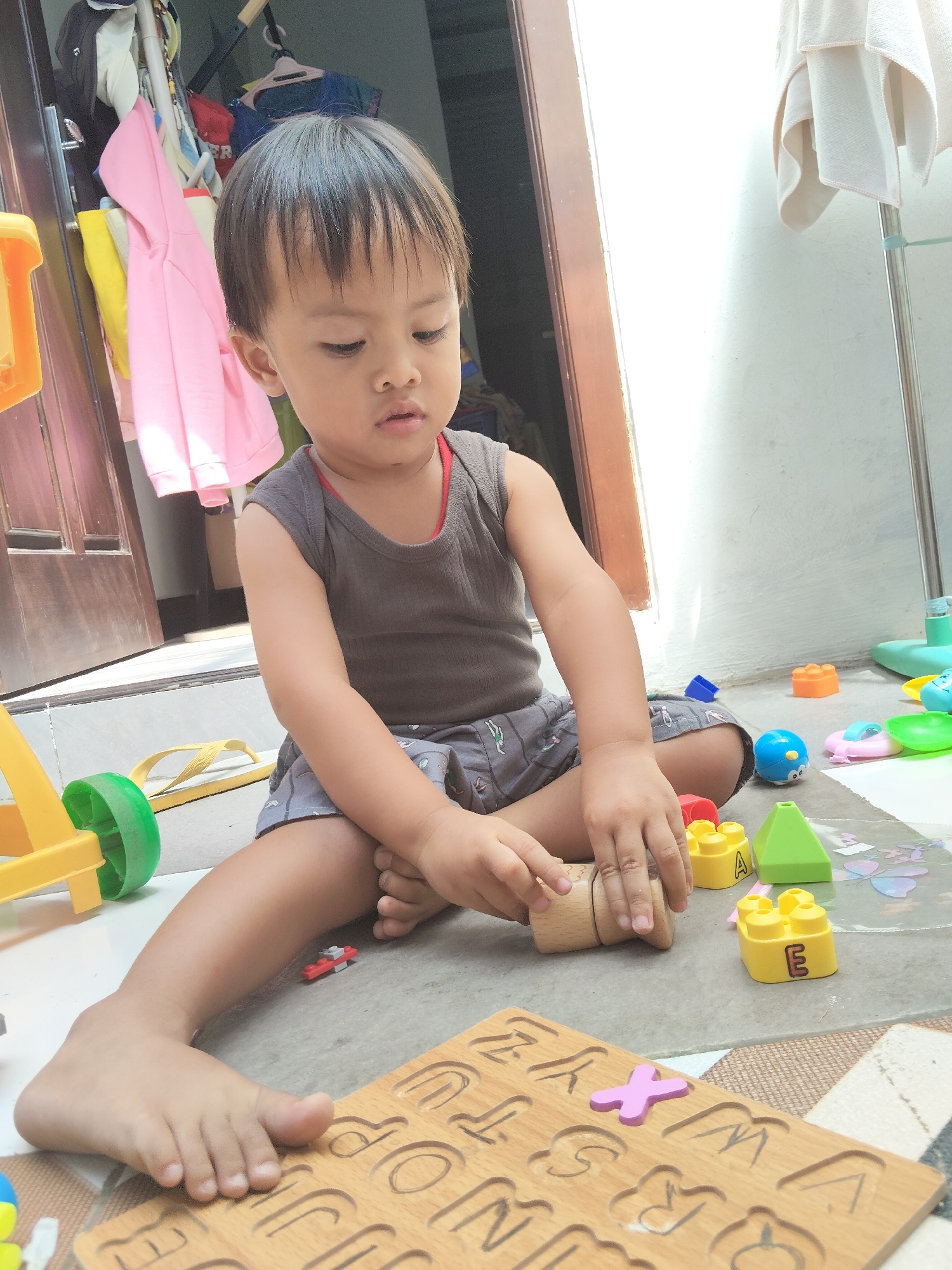 A young child, appearing to be of Southeast Asian descent, sits on a gray tiled floor in a shallow doorway, engaged with toys. The child has dark hair, is wearing a dark gray sleeveless shirt and gray patterned shorts, and is focusing intently on a wooden toy block they are holding. Scattered around the child are various colorful plastic toys, including building blocks with letters, a blue ball-shaped toy, and a green and yellow toy vehicle with a wheel. In the foreground, a wooden alphabet puzzle board with letters and some letter pieces is visible. Behind the child, a brown wooden door frame and a wall are present, with clothing hanging on a rack to the left. To the right, a light-colored towel is draped over a silver pole, and further in the background, a white wall is illuminated by sunlight. The scene suggests an outdoor or semi-outdoor area, possibly a residential space. The image is captured in Pemalang, Indonesia.