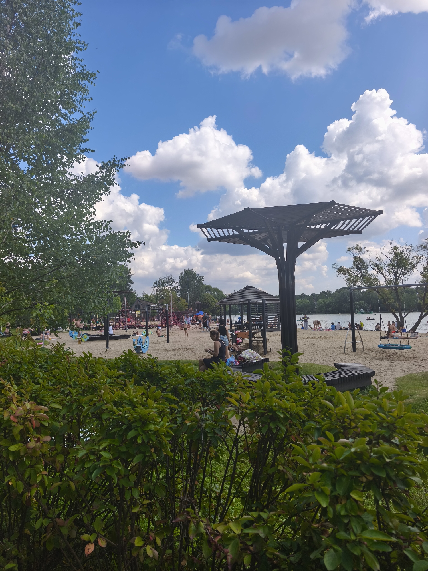 The image depicts a crowded beach scene on a sunny day with scattered clouds. In the foreground, dense green bushes obscure the lower portion of the scene. Beyond the bushes, a sandy beach stretches out, occupied by numerous individuals. Several children's playground structures, including swings and climbing frames, are visible on the sand. A large, dark wooden canopy structure stands prominently in the midground, offering shade. People are scattered across the beach, some sitting, some standing, and others appearing to be engaged in various activities. A body of water, likely a lake or river, forms the background, with a treeline visible on the far bank. Several small boats can be seen on the water. The overall impression is of a popular recreational area, likely a public beach or park, on a summer day. The location is identified as Moscow, Russia.