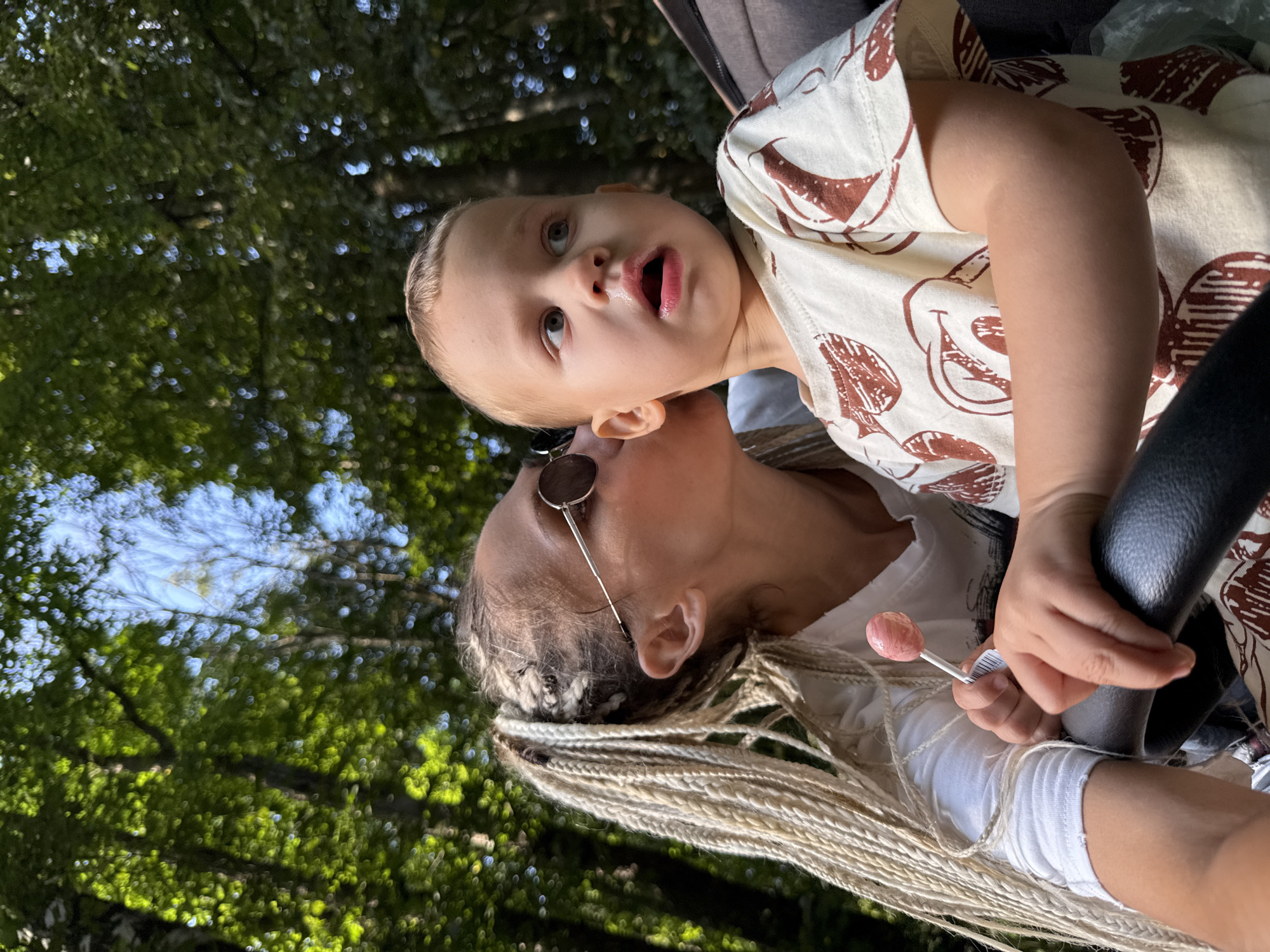 In Rusanovo, Russia, a medium shot captures a woman with blonde dreadlocks kissing a young child on the cheek. The woman wears sunglasses and a white t-shirt. The child, dressed in a light-colored shirt with a brown pattern, is seated in a stroller and has their mouth open. The child is holding a pink lollipop. They are outdoors, surrounded by lush green trees.