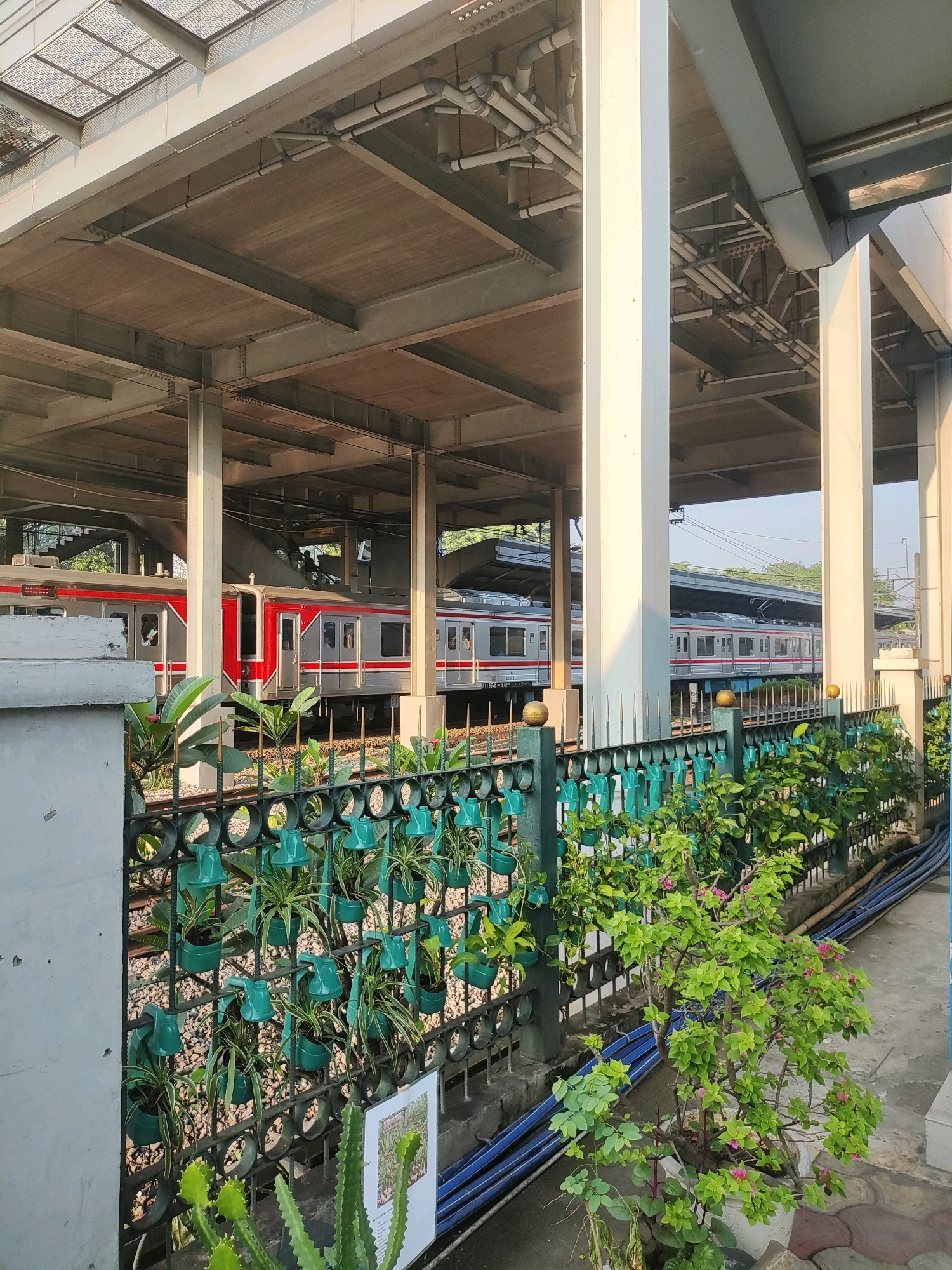 The image shows a train at a station in East Jakarta, Indonesia. The train is red and silver. There is a fence with plants in front of the train. The station has a covered roof supported by columns. The sky is clear and blue.