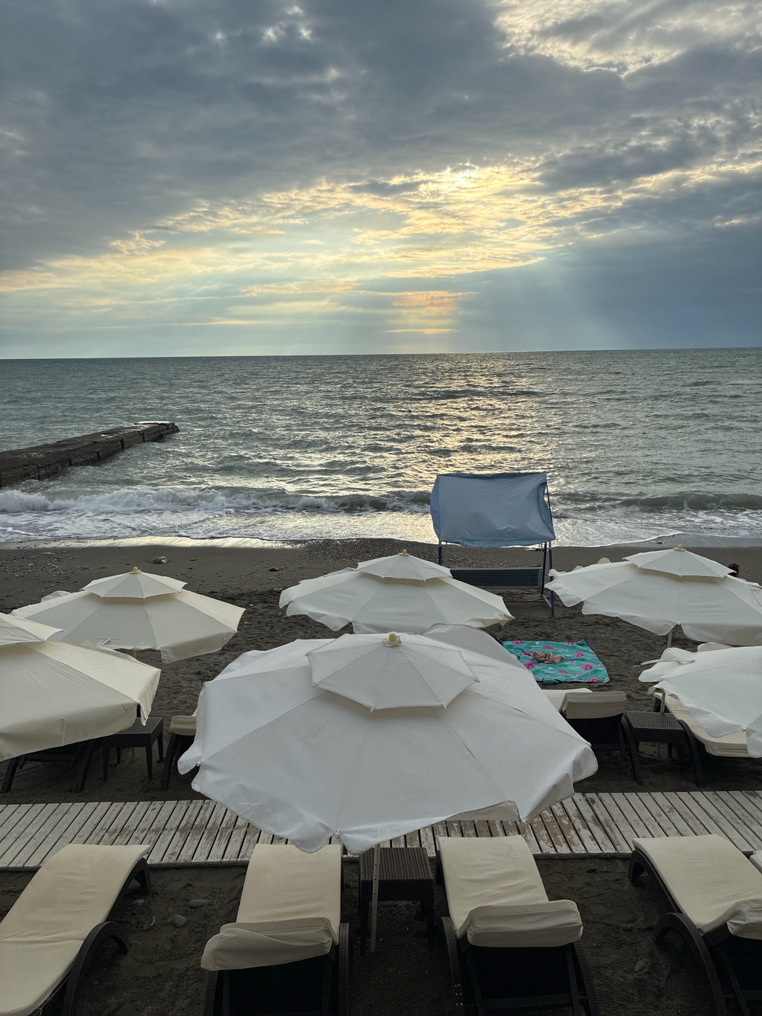 The image depicts a cloudy day at a beach in Sochi, Russia. In the foreground, several empty white beach umbrellas are open, providing shade over empty lounge chairs arranged on wooden planks. The sand is dark, and the sea is visible in the background, with moderate waves rolling in. A concrete pier extends into the sea on the left. On the right side of the beach, a blue screen or shelter stands near the water's edge. The sky is overcast, with breaks of sunlight filtering through the clouds, creating a dramatic effect over the sea. There are no people visible in the frame.