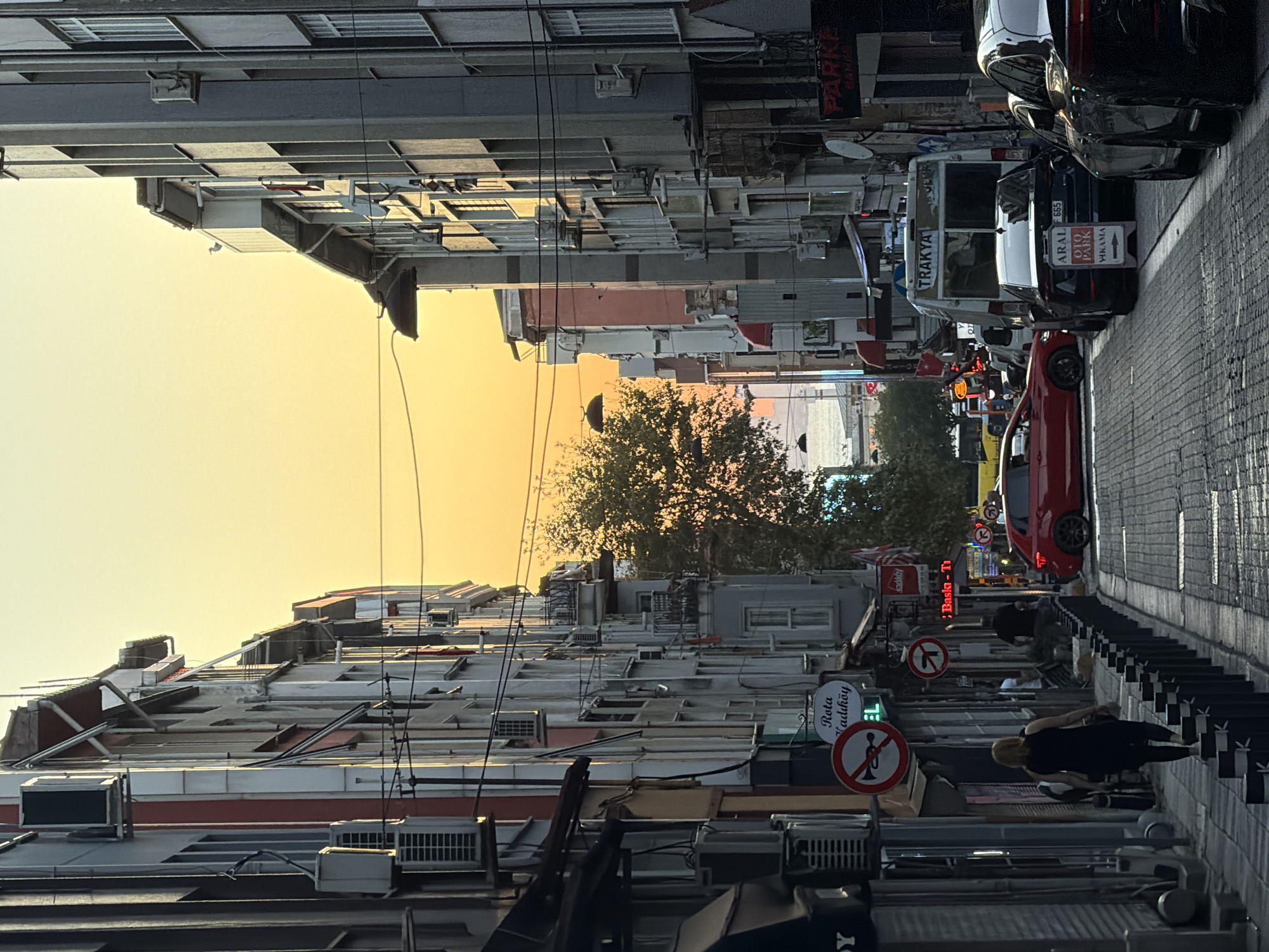 This image depicts a street scene in Kadıköy, Turkey, during sunset. Tall, narrow buildings line both sides of the cobblestone street, casting long shadows. The sky is a vibrant gradient of orange and yellow, with a few trees visible in the distance.
On the left side of the street, a woman in a black sleeveless dress is walking away from the camera, her back to the viewer. Several storefronts and air conditioning units are visible on the building facades. A circular red and white sign with a horn symbol indicates no honking. Another sign with the text "Rota Kadıköy" is also visible.
On the right side of the street, several cars are parked, including a red sedan in the middle of the road, a dark sedan with an "OTO PARK" sign, and a white van with "TRAKYA" written on its back. A large sign with "PARKE" is mounted on one of the buildings. The overall impression is a bustling urban environment bathed in the warm light of the setting sun.
