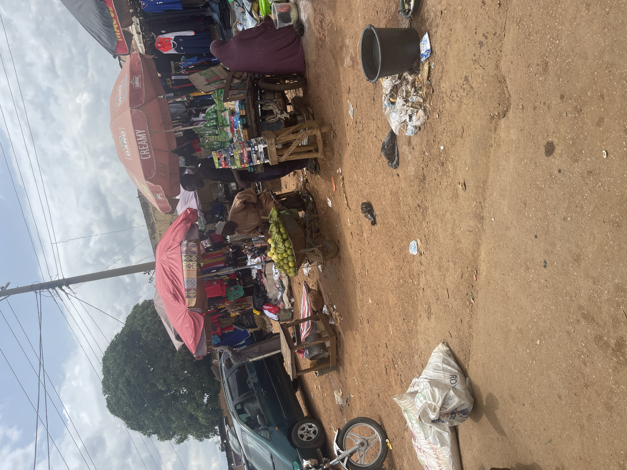 This image depicts an outdoor market scene in Rafin Par, Nigeria. A green minivan is parked on the left side of the frame, and a motorcycle is partially visible in the foreground. Several market stalls are set up, covered by umbrellas, displaying various goods including clothing and produce. Two men are visible behind a cart filled with green bananas. A woman in a purple headscarf sits on the right side of the frame, near a large black bucket and a pile of trash. The ground is dirt, with scattered debris. The sky is overcast.