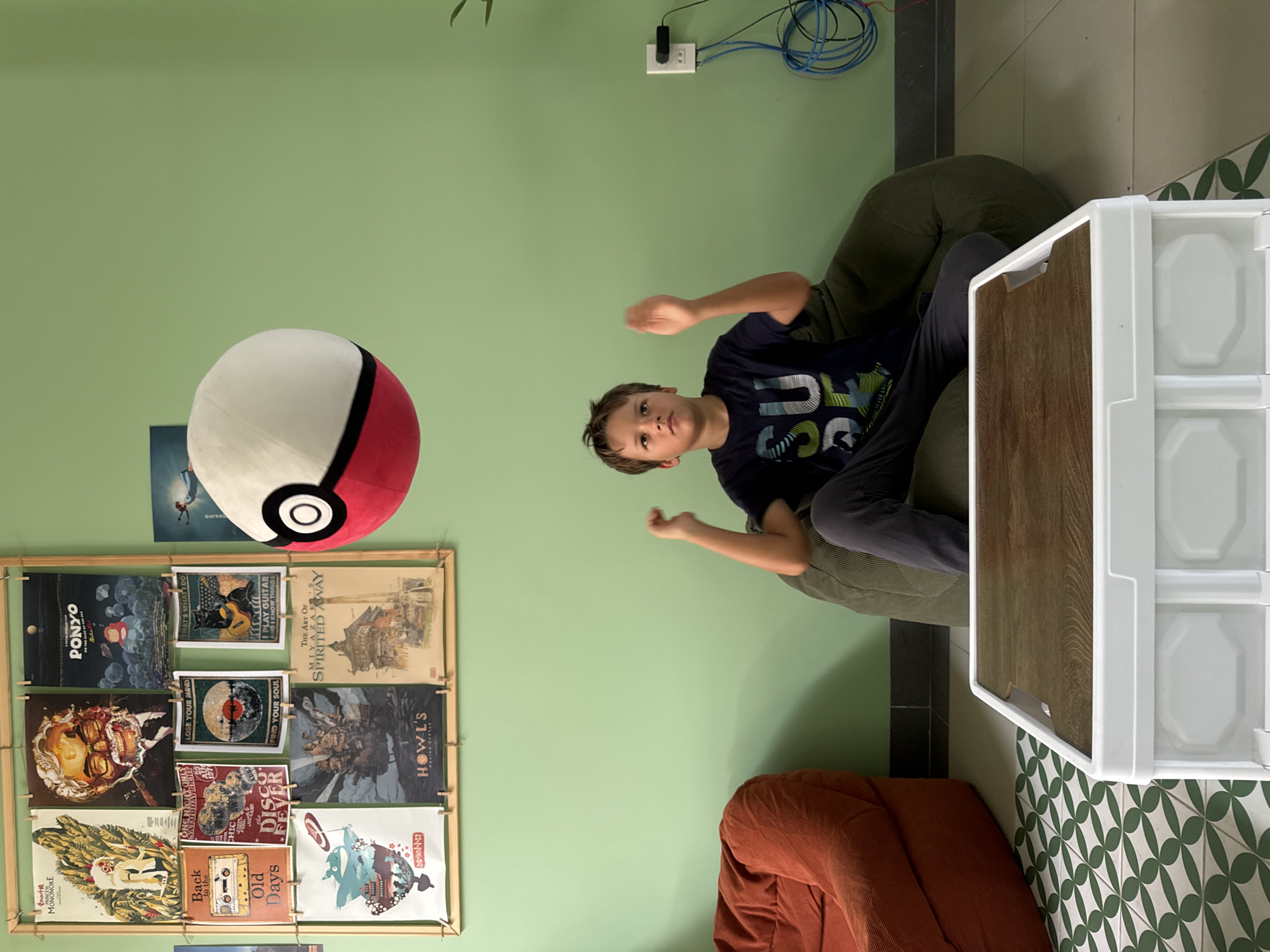 The image presents a rotated view of an interior setting in Phu Thuy Ward, Vietnam. The main subject is a child with short brown hair, wearing a navy blue t-shirt with graphic designs, seated in a large, olive-green beanbag chair. The child is positioned near a white cabinet with a faux wood-grain door. Above and to the left of the child is a large, plush Poké Ball decoration hanging against a pale green wall.
Several framed posters hang on the wall, featuring titles like "Ponyo," "Spirited Away," "Howl's Moving Castle," and "Princess Mononoke," along with other music and film-related artworks.
The floor is tiled with a green and white patterned tile. An orange beanbag chair is also visible in the bottom corner. Electrical wiring and an outlet are positioned on the wall near the upper edge of the frame. The overall lighting in the room is bright and appears to be natural.