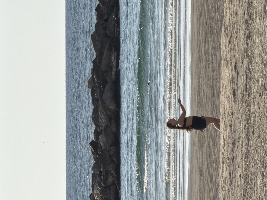 A female subject is depicted in profile, facing left, on a sandy beach in Tel-Aviv, Israel. She has long dark hair, possibly in a braid or ponytail, and is wearing a dark bikini top and dark shorts. Her right arm is extended forward, holding a dark, flat beach paddle. Her left leg is bent at the knee with the foot slightly raised, indicating a walking motion. In the mid-ground, the Mediterranean Sea shows small waves breaking on the shore. A dark rock breakwater spans horizontally across the water further out. A small, dark, spherical object is visible floating in the sea between the individual and the breakwater. The background consists of the blue sea meeting a clear, light-toned sky at the horizon.
