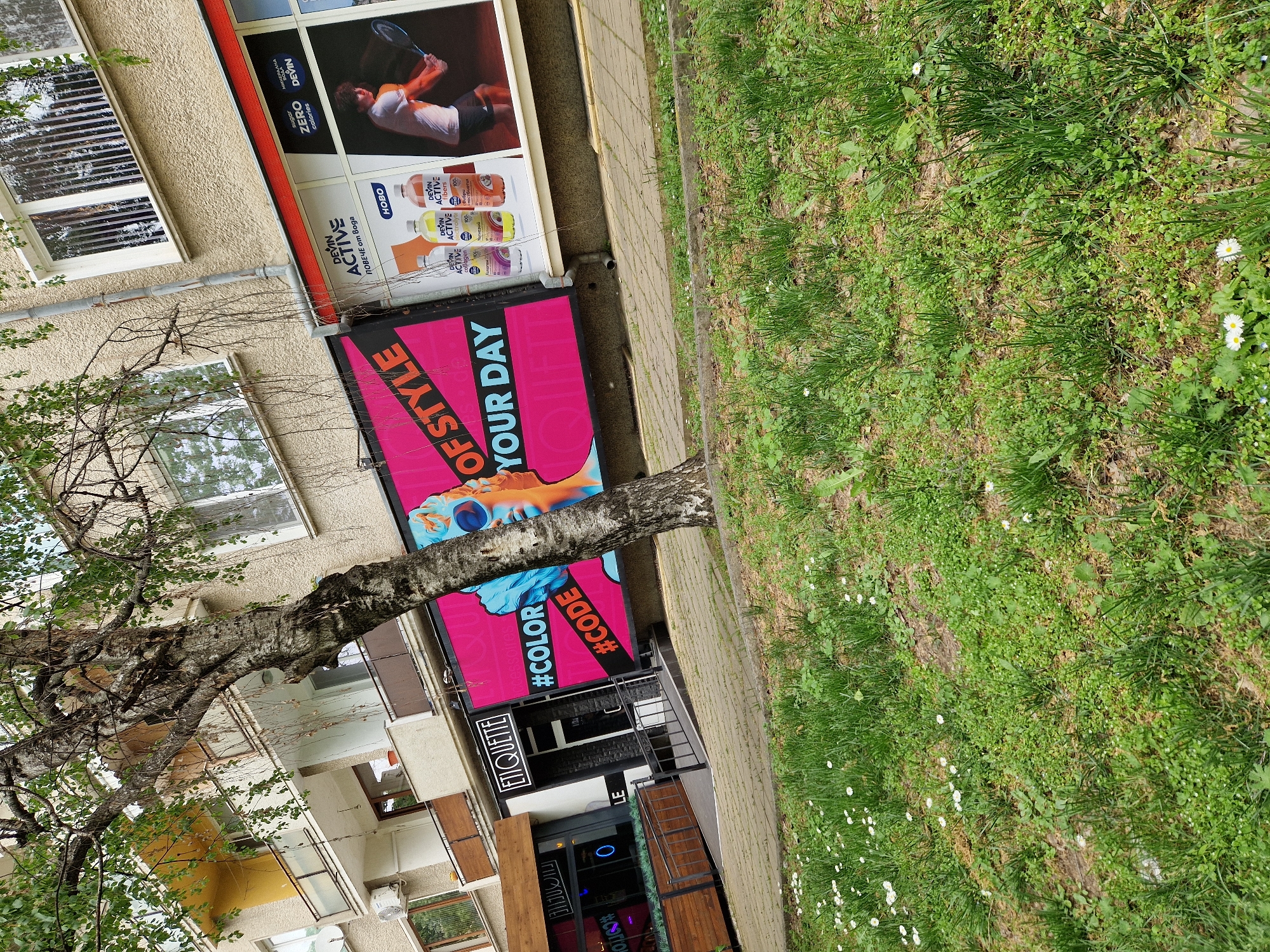 The image shows a street scene in Lovech, Bulgaria, with a multi-story building dominating the left side of the frame. The building features several windows and balconies, along with advertisements for "Devin Active" and "ETIQUETTE" which include hashtags such as "#COLOR," "#CODE," and phrases like "OF STYLE" and "YOUR DAY." A tree with sparse foliage extends from the foreground, partially obscuring the building. To the right of the building is a paved sidewalk and a patch of green grass interspersed with white daisies. The scene is captured in daylight, presenting a typical urban landscape.