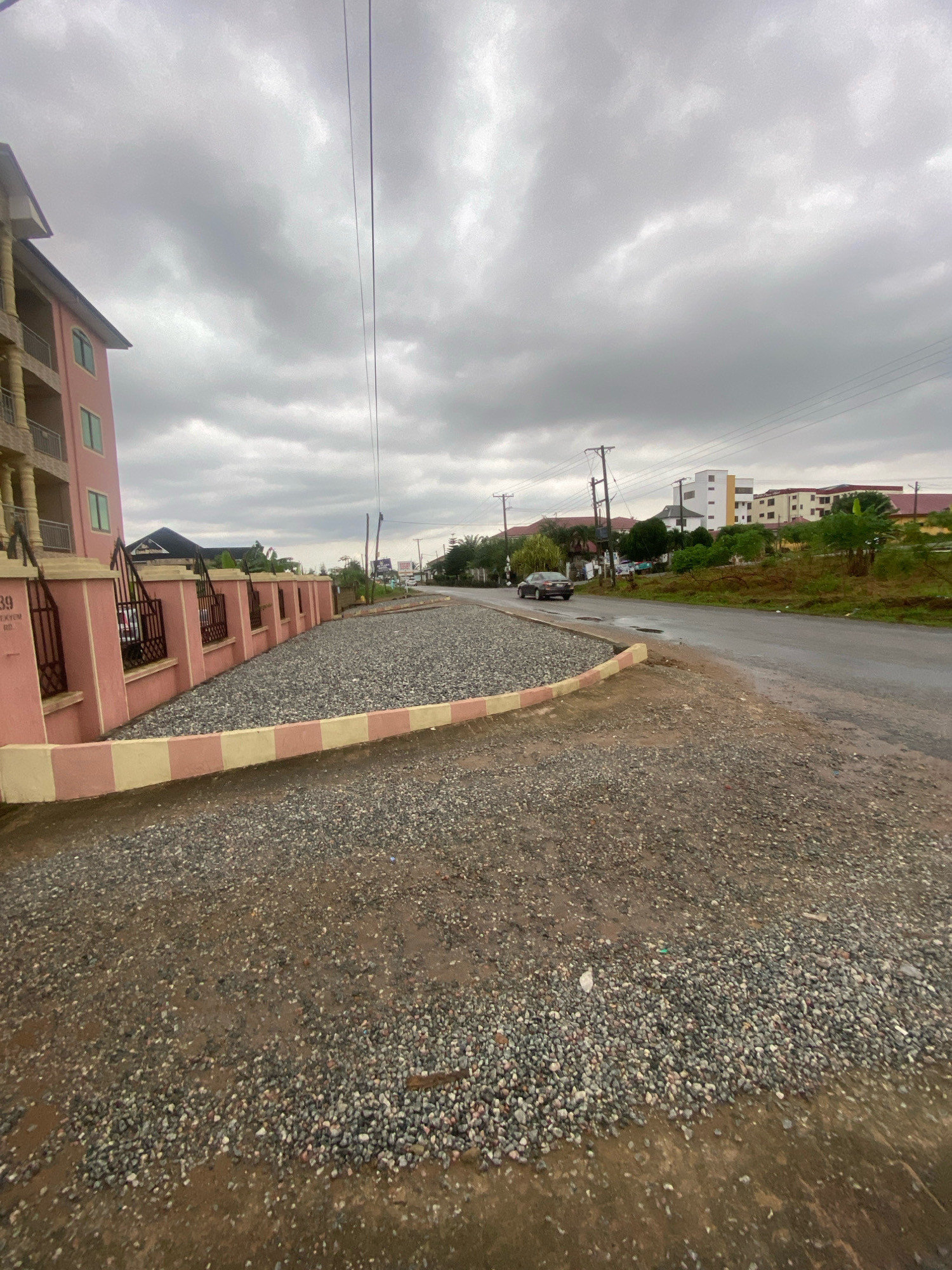 The image depicts a street scene in Koforidua, Ghana. On the left side of the frame, a pink building with multiple stories and balconies is visible. A low wall with iron fencing runs along the front of the property, leading to a gravel-covered area. The wall is decorated with bands of pink and yellow. A road runs parallel to the wall, with a car driving away from the viewer. Beyond the road, there are several other buildings of varying sizes and colors, some with red roofs, and lush green vegetation. Utility poles with electrical wires are present throughout the scene. The sky is overcast with heavy gray clouds, suggesting a possibly rainy or dull day. The ground in the foreground is also covered in gravel and appears somewhat wet.