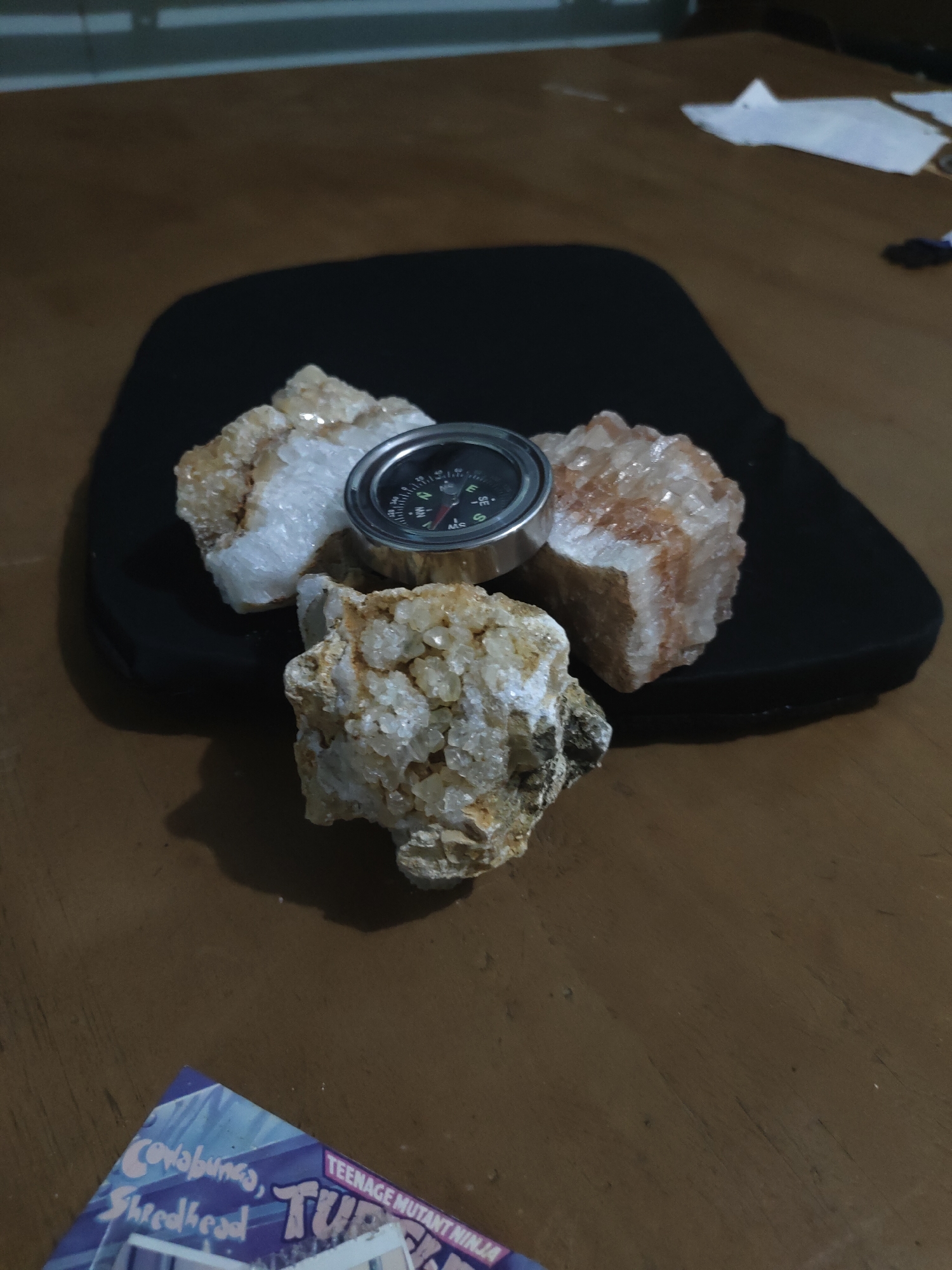 The image displays a still life composition of three mineral clusters and a small metallic compass resting on a dark, irregularly shaped pad, all situated on a brown wooden table.
The central subject is a silver-rimmed compass with a black face, featuring white cardinal directions (N, E, S, W) and degree markings. Its red needle points roughly towards the top-left of the frame. Surrounding the compass are three distinct mineral clusters, likely quartz, each exhibiting numerous small, somewhat translucent crystals. The cluster positioned at the top-left appears predominantly white with some yellowish-brown tones, while the top-right cluster shows more pronounced reddish-orange coloration mixed with white crystals. The cluster at the bottom is rougher, displaying clear crystals embedded in a mottled, earthy-toned matrix. These items are arranged in a triangular formation on a matte black pad.
The surface beneath is a medium-brown wooden table with visible grain running horizontally, indicating natural wood. In the bottom-left foreground, a partial view of a colorful magazine or comic book cover is visible. Identifiable text includes "Cowabunga," "Shredhead," "TEENAGE MUTANT NINJA," and "TUR," suggesting a Teenage Mutant Ninja Turtles publication.
The background elements are out of focus. To the upper-left, a blurry brown surface suggests a wall or furniture. To the upper-right, indistinct white crumpled objects, possibly paper or tissue, are visible on a similar brown surface, along with a small, dark, elongated object. The uppermost part of the image shows a horizontal, light-colored strip, possibly a window sill or architectural detail.
The scene is lit with what appears to be ambient or indirect light, creating soft shadows and highlights on the crystalline surfaces and the compass rim.
The specific location within Davao City, Philippines, cannot be determined from the visual evidence presented in the image.