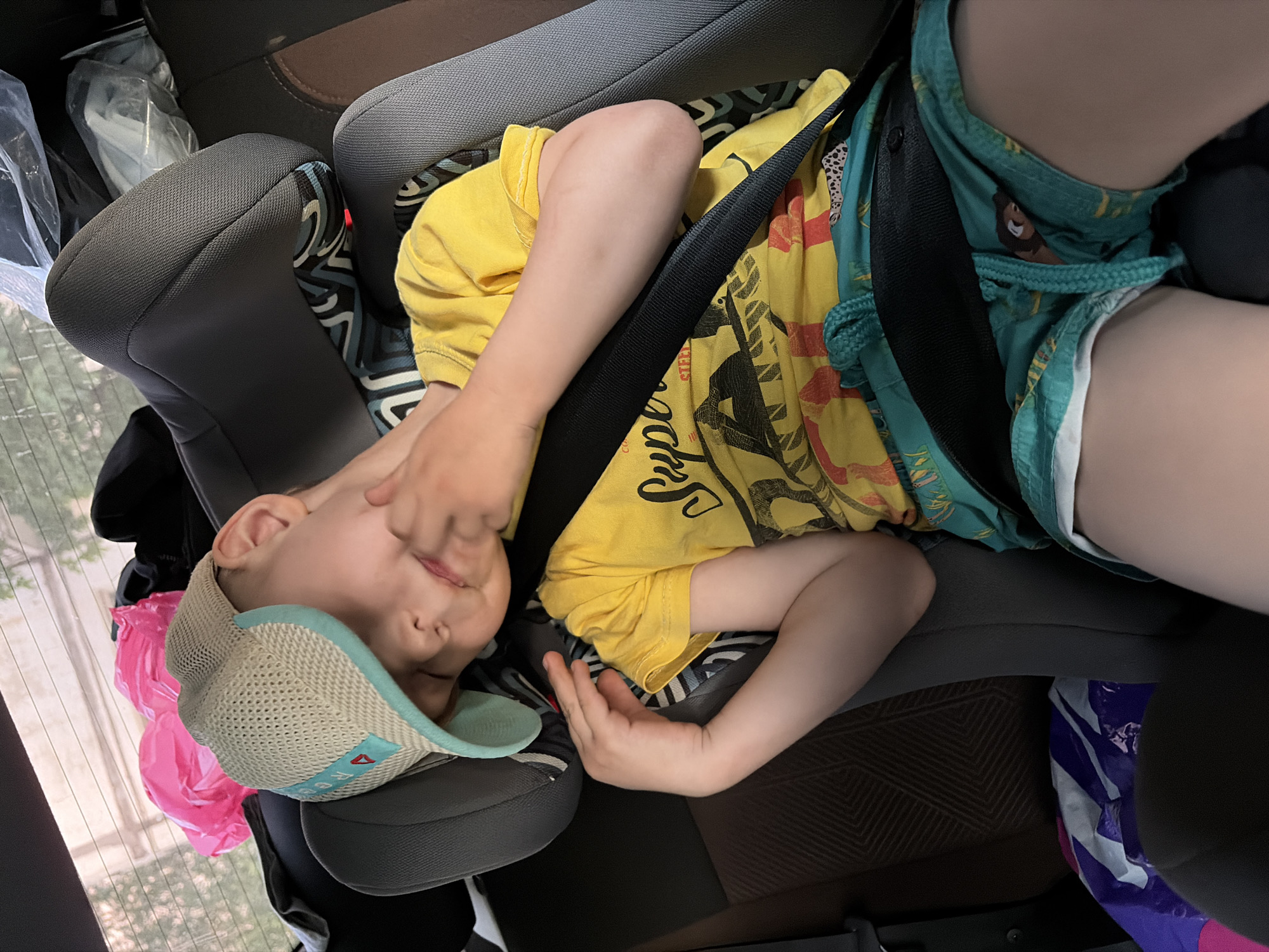 A child is asleep in a car seat. The child is wearing a yellow t-shirt, green shorts, and a baseball cap. The child is secured in the car seat with a black seatbelt. The location cannot be determined from the image alone.