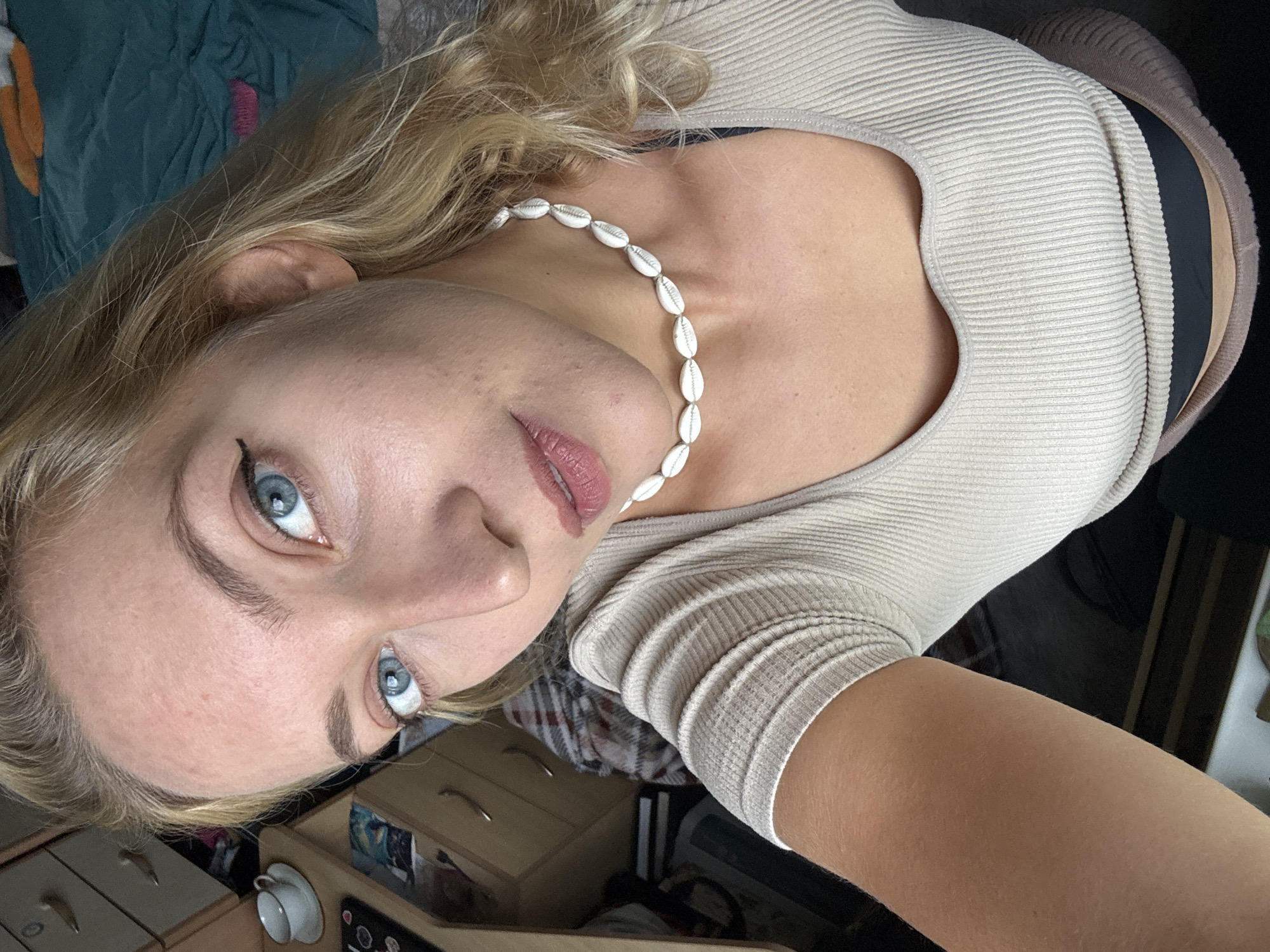 The image shows a fair-skinned blonde woman with blue eyes wearing a beige ribbed top and a seashell necklace. She is looking upwards towards the camera. Part of a brown skirt and a black undergarment are visible near her waist. The background includes a green and orange blanket, a wooden dresser, and what appears to be a cup on a small table. There is no information indicating that this image was taken in Minsk, Belarus.