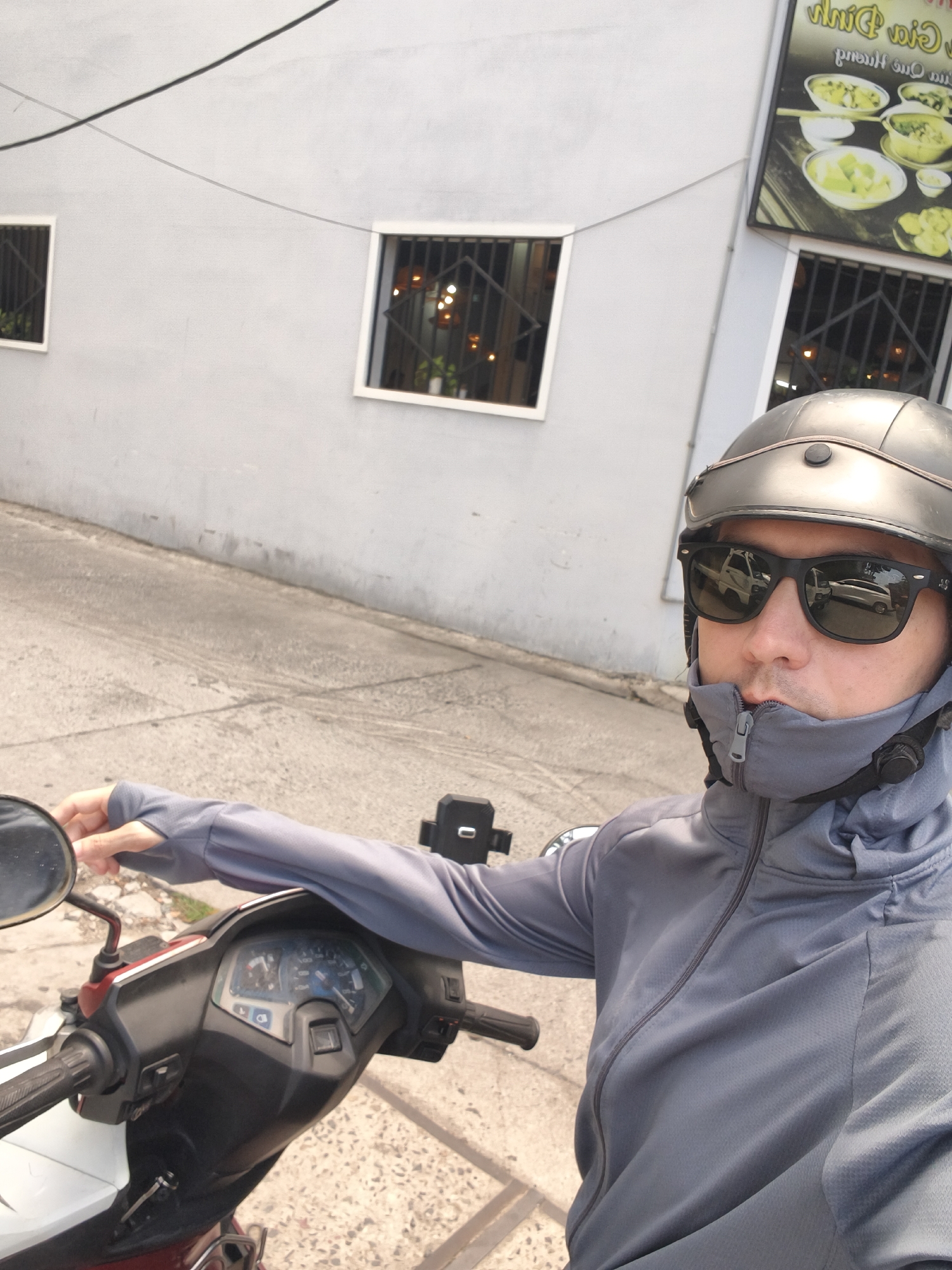 An individual is depicted in a selfie, seated on a white and dark red scooter, holding the handlebars. The person wears a dark grey helmet, black sunglasses, and a grey full-zip jacket with long sleeves that includes a high collar or integrated neck covering. The sunglasses reflect a white truck and another vehicle.
The setting is outdoors, likely a street or alleyway, with a light grey building in the background. The building features two square windows secured with black metal grates. To the right, a partially visible vertical sign advertises food, displaying images of bowls filled with a light-colored dish and some fried items. Visible text on the sign includes "Phở Nồi Đất" (Clay Pot Pho) and "Bánh," along with other Vietnamese characters. Two dark electrical wires are strung across the upper portion of the background.
The ground is concrete, showing signs of wear and cracks. The bright lighting suggests it is daytime. The person appears to be pausing during or before a scooter ride. The type of scooter, the architectural details, and the Vietnamese text on the food sign are consistent with the provided location context of Bac Nha Trang Ward, Vietnam.