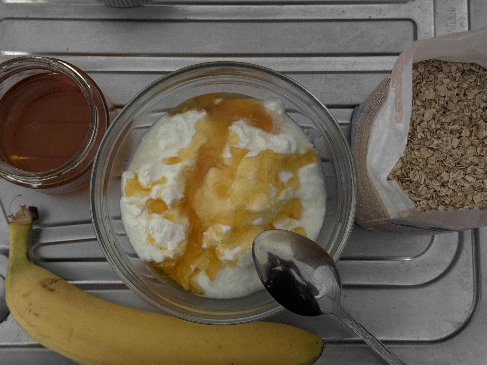 This overhead shot displays a collection of breakfast or snack ingredients arranged on a metallic, ridged surface, possibly a kitchen drainer or tray. In the center, a clear glass bowl holds a creamy white substance, likely yogurt or cottage cheese, generously drizzled with golden honey, creating an inviting visual. To the left of the bowl, a glass jar brimmed with more amber honey is visible. A partially opened paper bag filled with rolled oats sits to the right of the bowl, its contents ready for use. A ripe yellow banana curves across the bottom of the frame, below the bowl. A silver spoon rests in the lower right, with the words "STAINLESS STEEL" etched onto its handle. The metallic surface features horizontal ridges and appears well-used. Faint letters "PYR" can be seen on the metallic surface in the upper right corner, possibly part of a brand name. The overall impression is one of simple, healthy food preparation in an indoor setting.