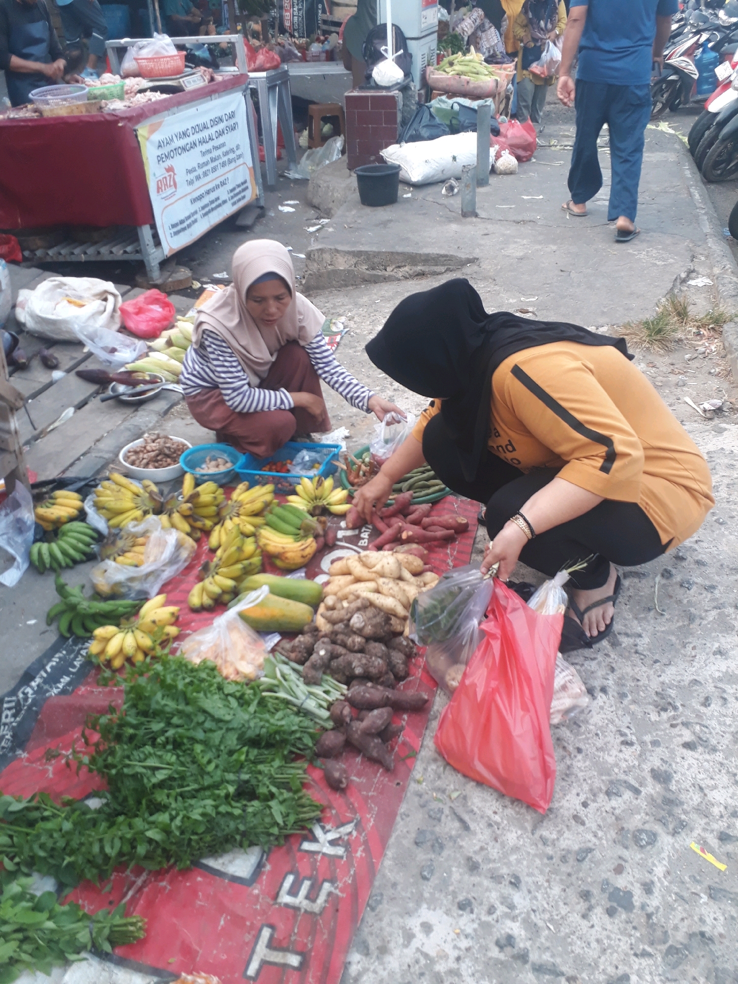 An outdoor marketplace in Pangkalpinang, Indonesia, features a woman wearing a light brown headscarf and a striped shirt, crouched on the ground amidst a variety of produce. She is sorting through a pile of sweet potatoes. To her right, another woman, wearing a black headscarf and an orange shirt, is also crouched and holding a red plastic bag filled with items. A variety of fruits and vegetables are displayed on a red mat on the ground, including bananas, green beans, and leafy greens. In the background, a food stall displays chicken, and a man in a blue shirt and pants walks away from the camera. Motorcycles are parked on the right side of the image, and the ground is paved concrete.