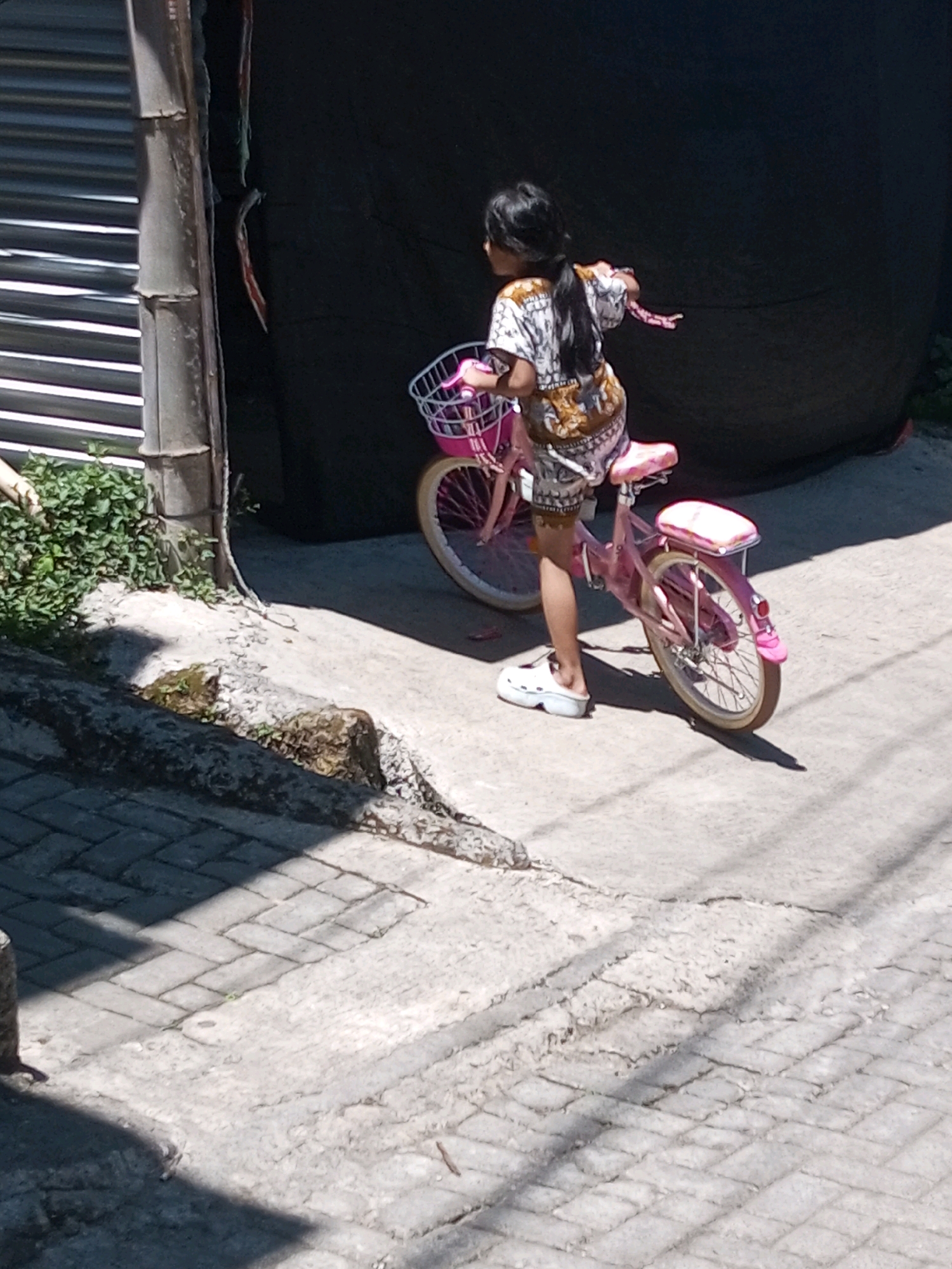 A young girl with dark hair tied in a ponytail rides a pink bicycle with a basket in front. She is wearing a patterned shirt and shorts, and white slip-on shoes. The bicycle has a pink seat and streamers hanging from the handlebars. The girl is on a paved sidewalk, with a dark wall or curtain behind her. To the left, there is a building with a corrugated metal facade and a drainpipe. Green foliage grows near the base of the building. The ground is cracked concrete and brick paving. The image was taken in Andongsili, Indonesia, on a sunny day, as evidenced by the shadows.