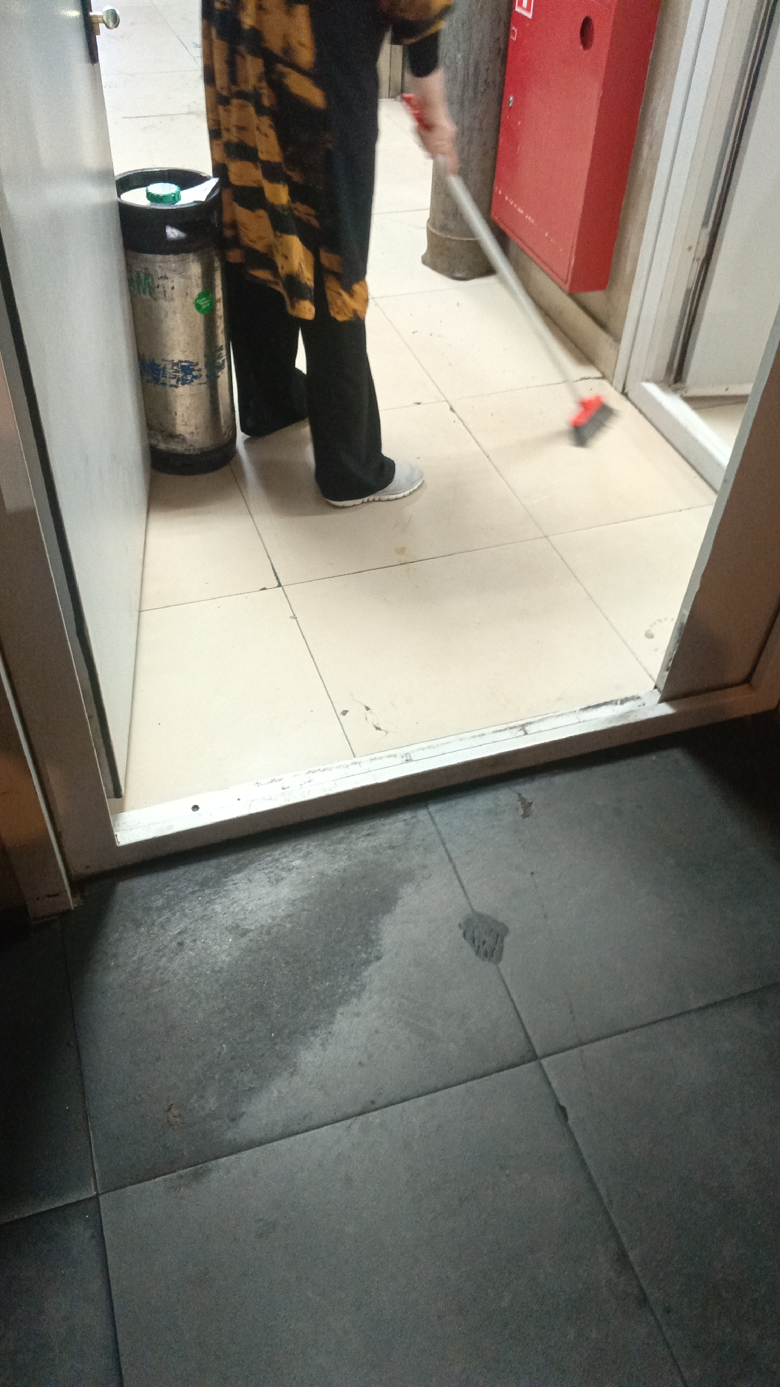 The image shows a person sweeping the floor inside what appears to be a public space, possibly an entryway or hallway. The person is wearing black pants and a long, dark tunic with a yellow and black patterned design. They are also wearing light-colored athletic shoes. The person is holding a broom with a red head and is actively sweeping the floor, which is made of light-colored square tiles. To the left of the person is a metal trash can with a green lid. In the background, to the right, there is a bright red fire alarm box mounted on a light-colored wall. The floor in the foreground, beyond the tiled area, is made of dark gray square tiles and shows signs of wetness or a spill. The overall scene suggests a cleaning or maintenance activity taking place in a building. There is no specific indication in the image that would place this scene in Mytishchi, Russia.