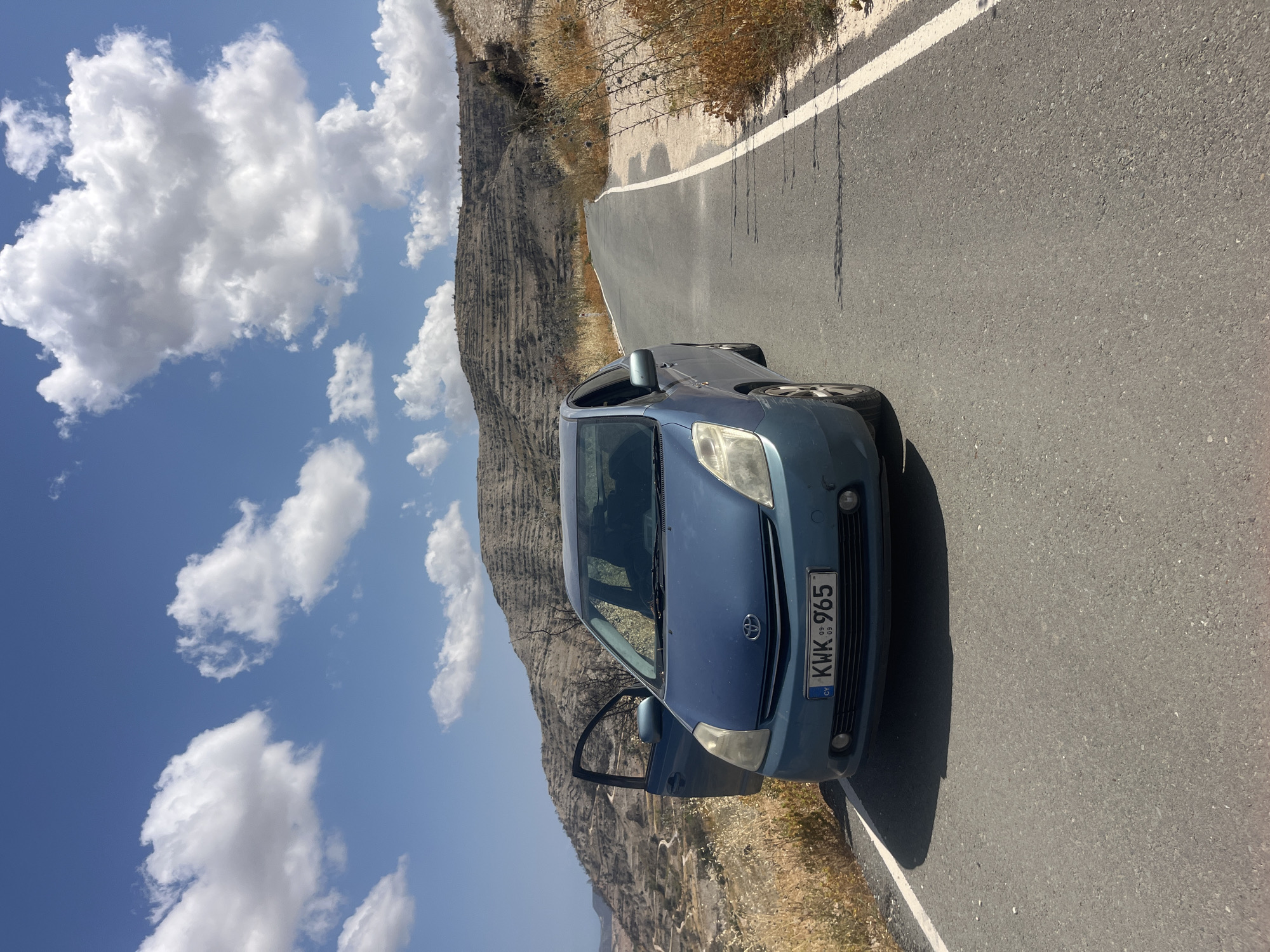 A blue Toyota Prius is parked on the side of a paved road in Agios Georgios Lemesou, Cyprus. The car's driver-side door is open. The road curves to the right and ascends a hill. The landscape is arid with dry grass and sparse vegetation on either side of the road. In the background, a barren hillside is terraced with numerous horizontal lines, suggesting past agricultural use. The sky is blue with scattered white clouds. The license plate of the car reads "KWK 965".
