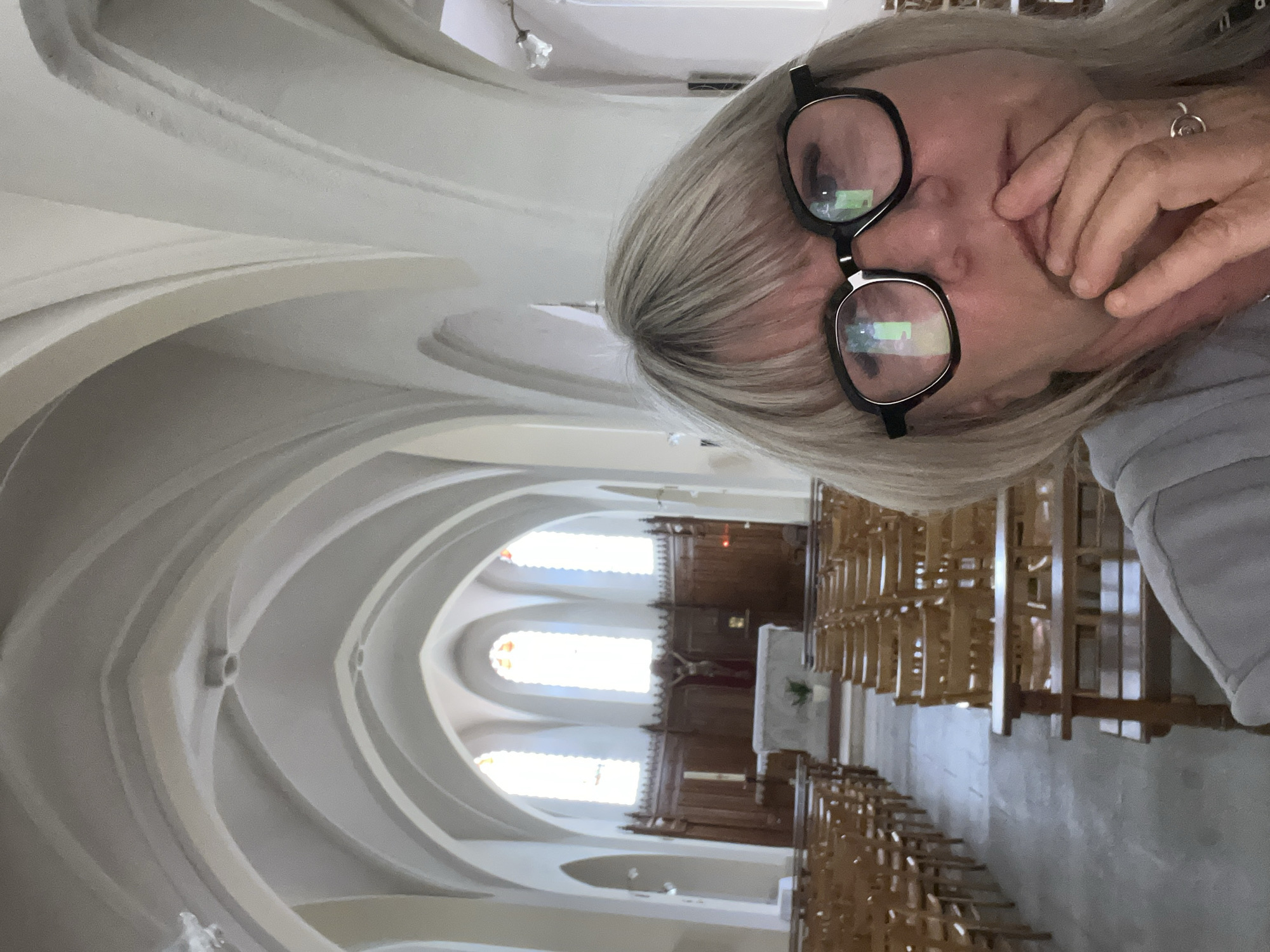 The image captures a woman with blonde hair and black-rimmed glasses in what appears to be the interior of a church. The woman is partially obscuring the view of the church's nave. The church features a vaulted ceiling with arches leading to an altar. Rows of wooden chairs line the aisle. The background suggests the church is located in Chambœuf, France.