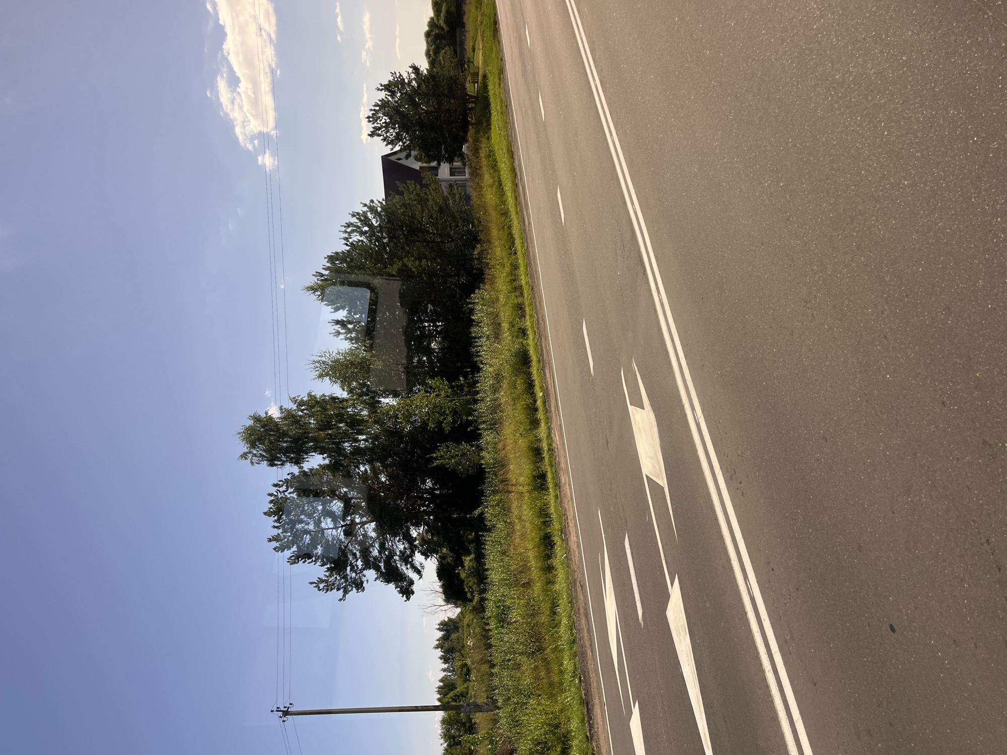 The image captures a roadside scene on a clear day. In the foreground, a paved road with white lane markings and directional arrows is visible, suggesting a straight stretch of highway. The road surface appears to be asphalt.
To the left of the road, a field of green grass and vegetation stretches towards a line of trees. A wooden utility pole with wires runs parallel to the road. Further in the background, a white house with a dark roof is nestled amongst the trees, indicating a rural or semi-rural setting. The sky is a clear blue with a few wispy clouds. The overall impression is one of a quiet, sunny day in the Belarusian countryside. The location is identified as Юнькаўскі сельскі Савет, Belarus. There are no people or animals visible in the frame.