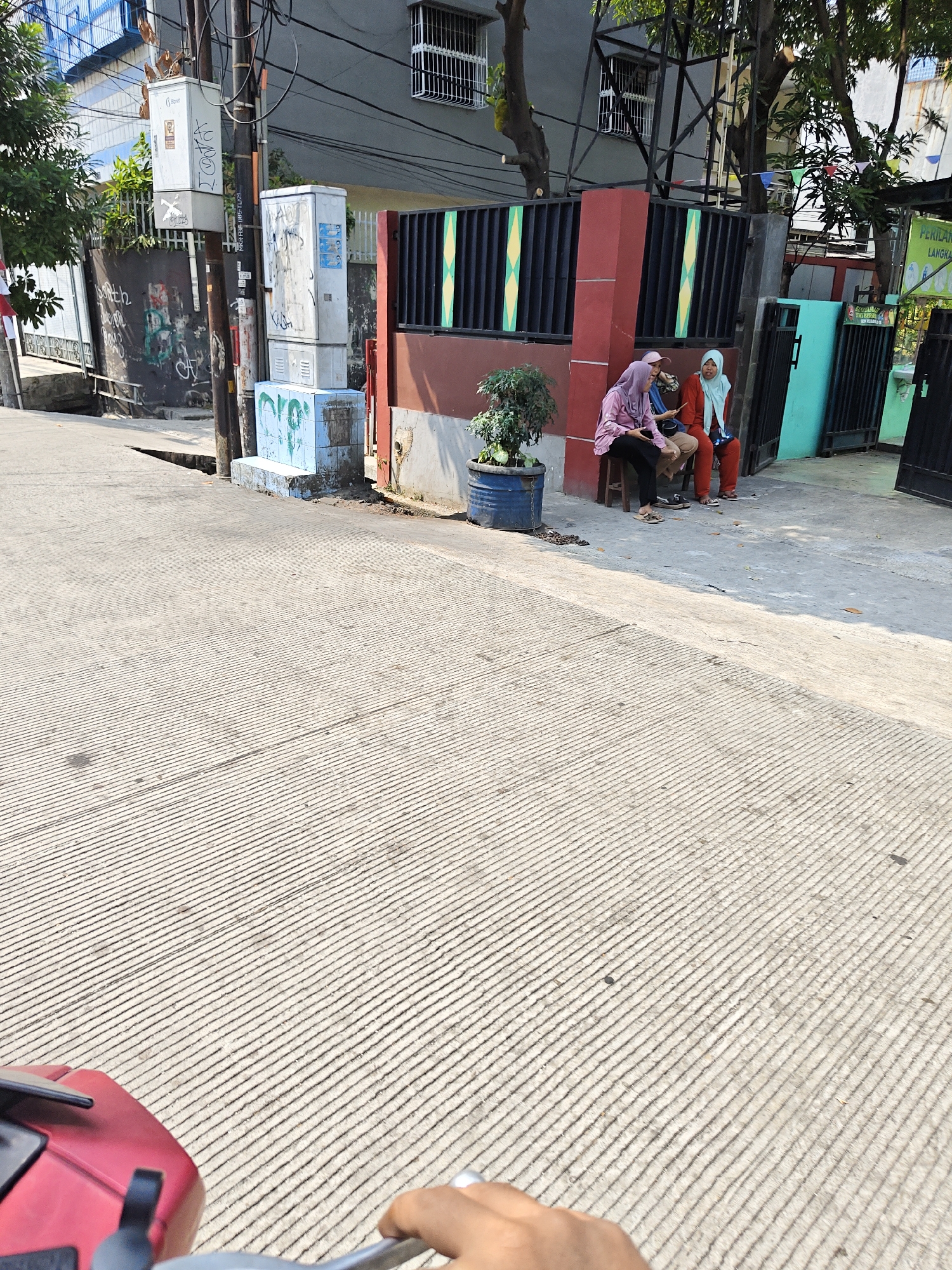 The image depicts a street scene in North Jakarta, Indonesia. In the foreground, a textured concrete road runs diagonally from the bottom left to the top right. A red scooter, partially visible at the bottom left, suggests a mode of transportation. In the middle ground, three individuals are seated on a low bench or ledge outside a building. Two women are wearing hijabs; one in a purple hijab and pink top, and the other in a light blue hijab and orange top. A third individual, whose face is partially obscured, is seated between them. A potted plant in a blue drum is placed to their left. Behind them, a building with a dark brown lower facade and dark metal fencing is visible. Electrical boxes and poles are prominent features along the side of the road, with one box covered in graffiti. Trees with green foliage are scattered throughout the scene, providing shade. The overall impression is an outdoor urban environment on a sunny day.